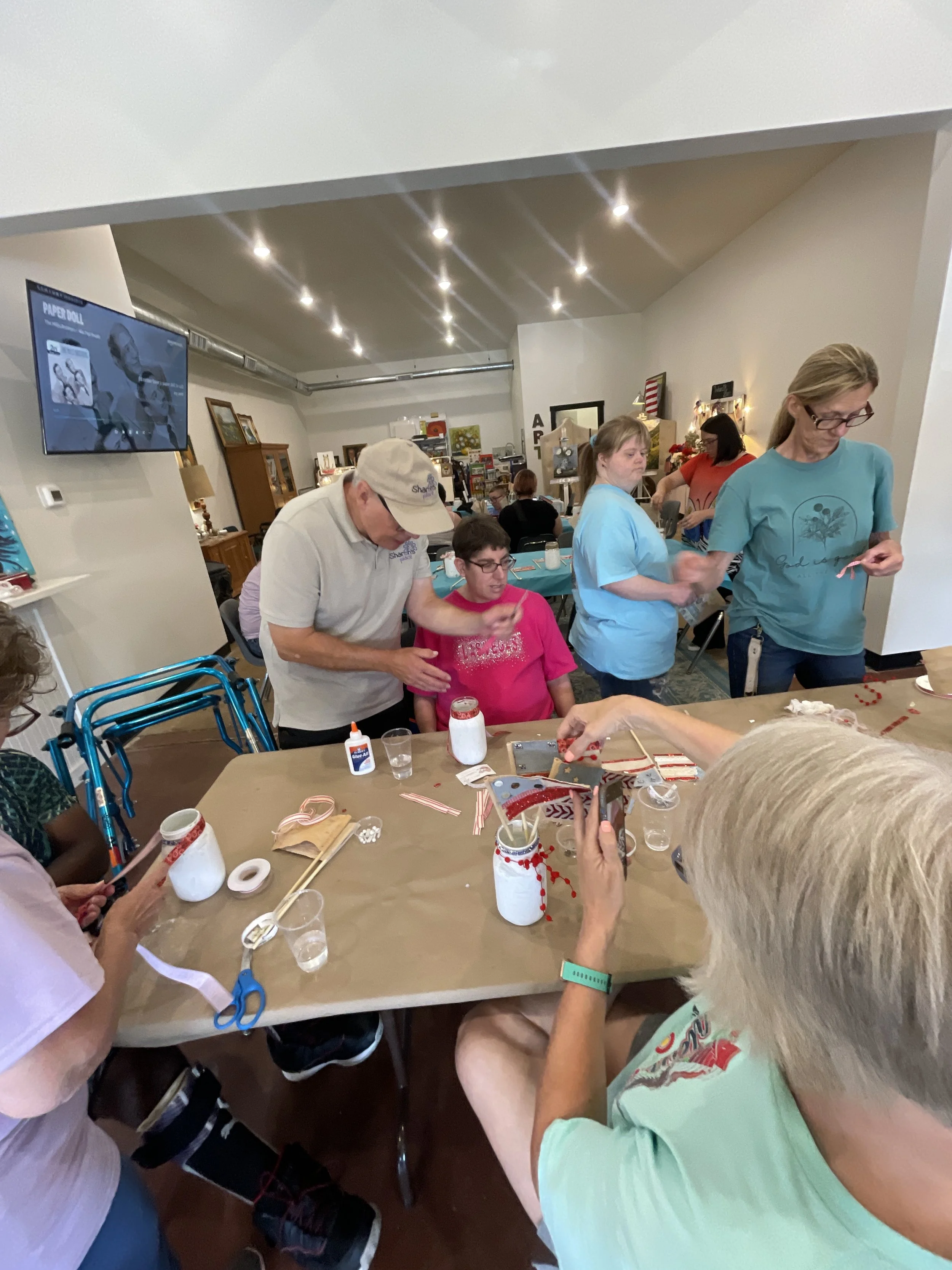 Group of people gathered around a table making crafts, with decorations including jars, ribbons, and glue, in a well-lit room.