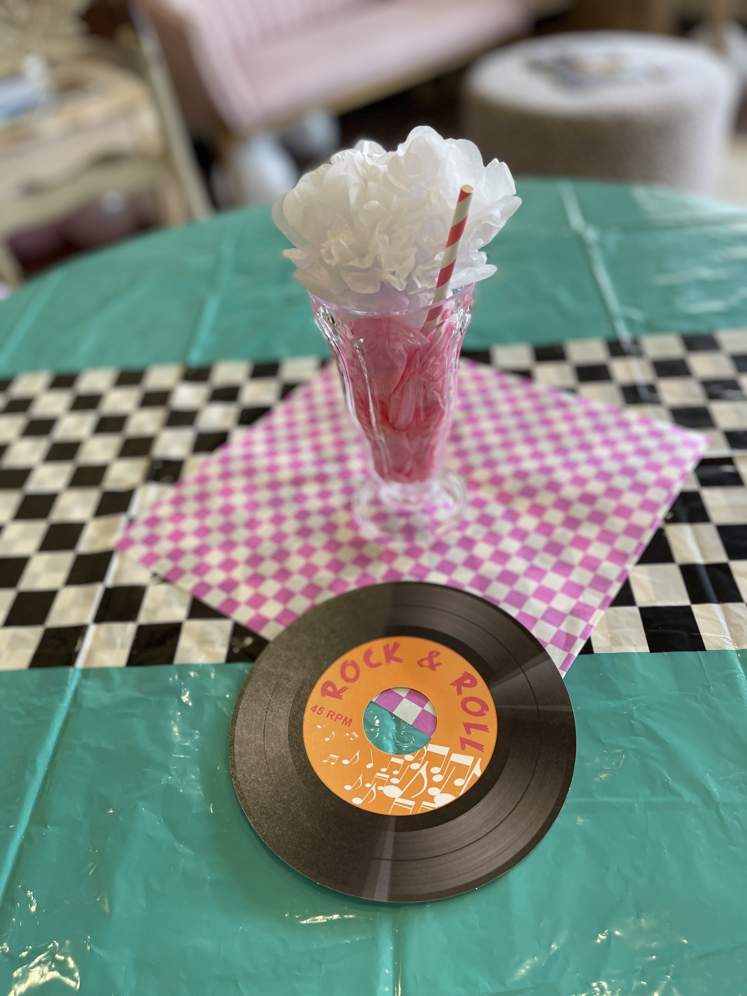 A decorative pink and white checkered napkin with a clear glass vase filled with pink and white tissue paper, a striped straw, and a black vinyl record with a colorful label reading 'Rock & Roll'.