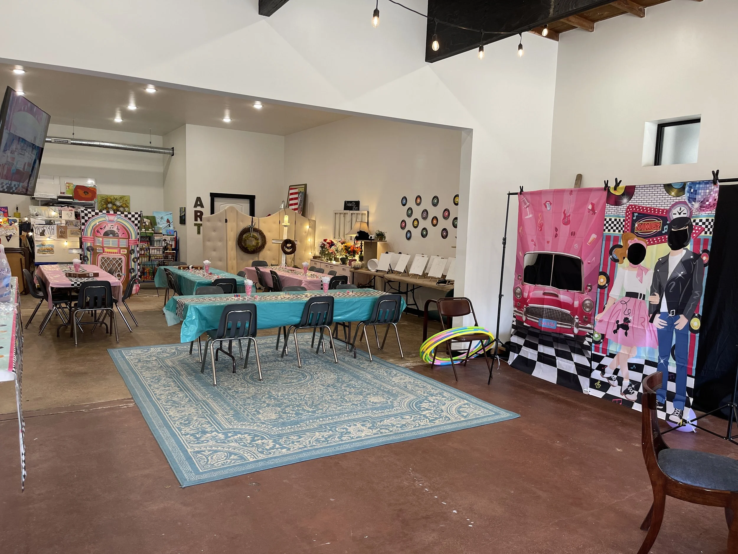 Decorated indoor space with tables covered in pink and teal tablecloths, framed photo booth with pink car backdrop, and a wall display of vinyl records.