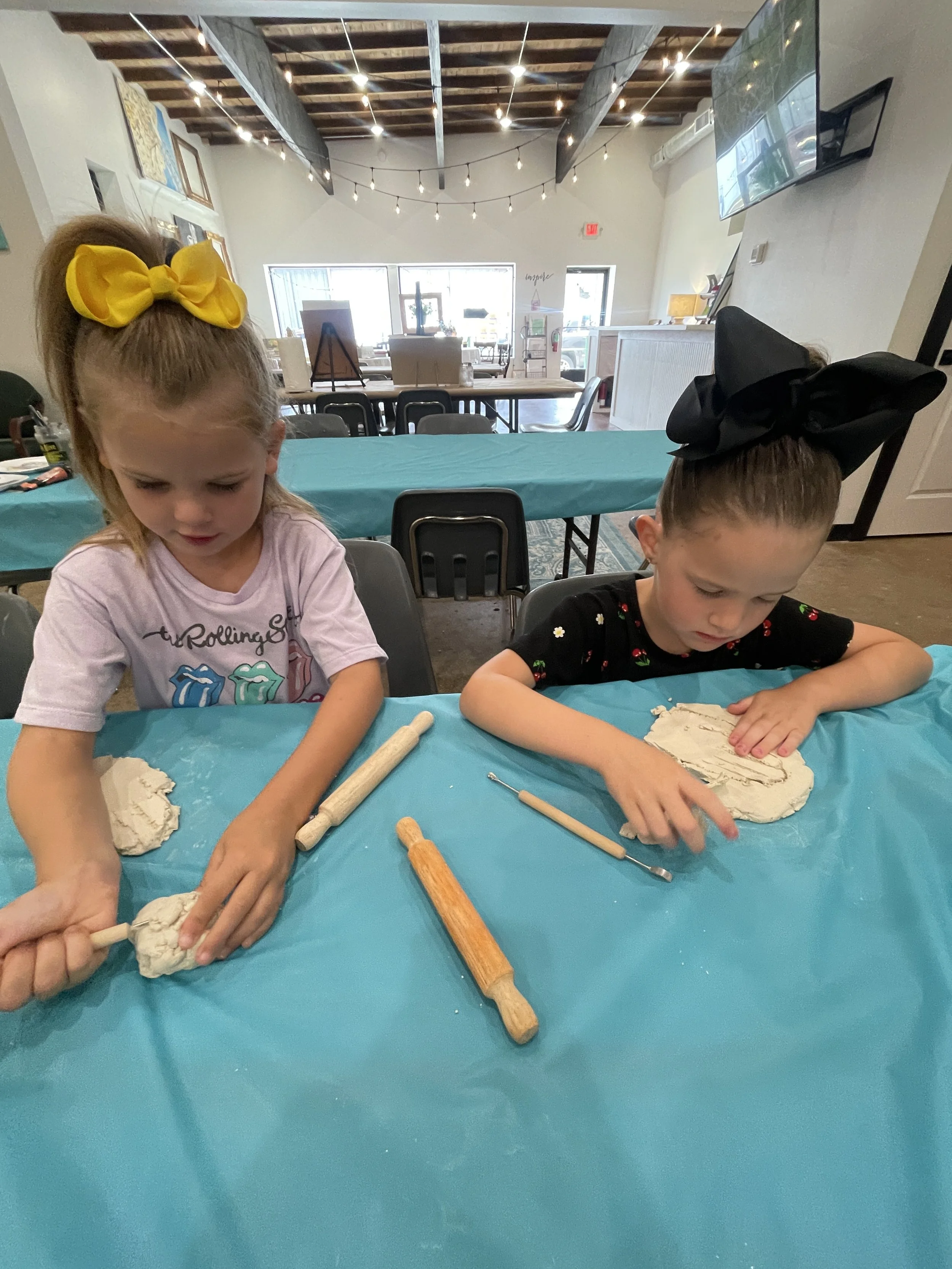 Two young girls are sitting at a table, rolling out clay with wooden tools. One girl has a yellow bow in her hair, and the other has a black bow. The table is covered with a blue tablecloth, and there are additional rolling pins on the table. In the