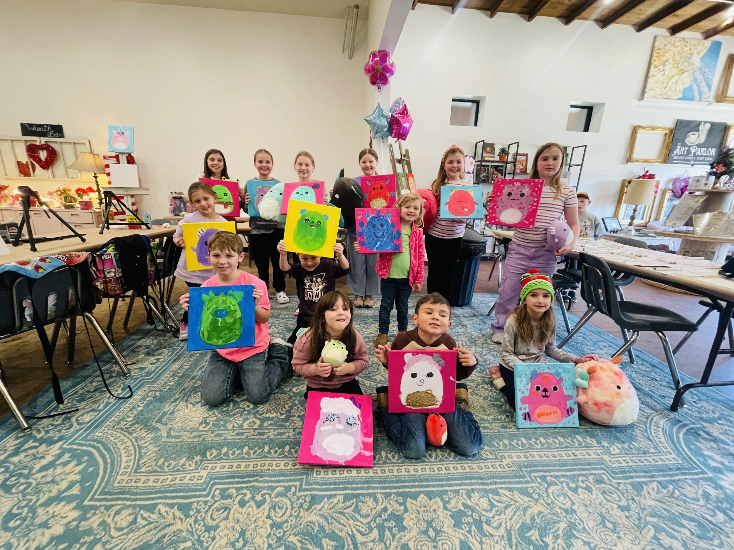 Children holding colorful, plush animal paintings in a classroom or art studio with balloons and art supplies in the background.