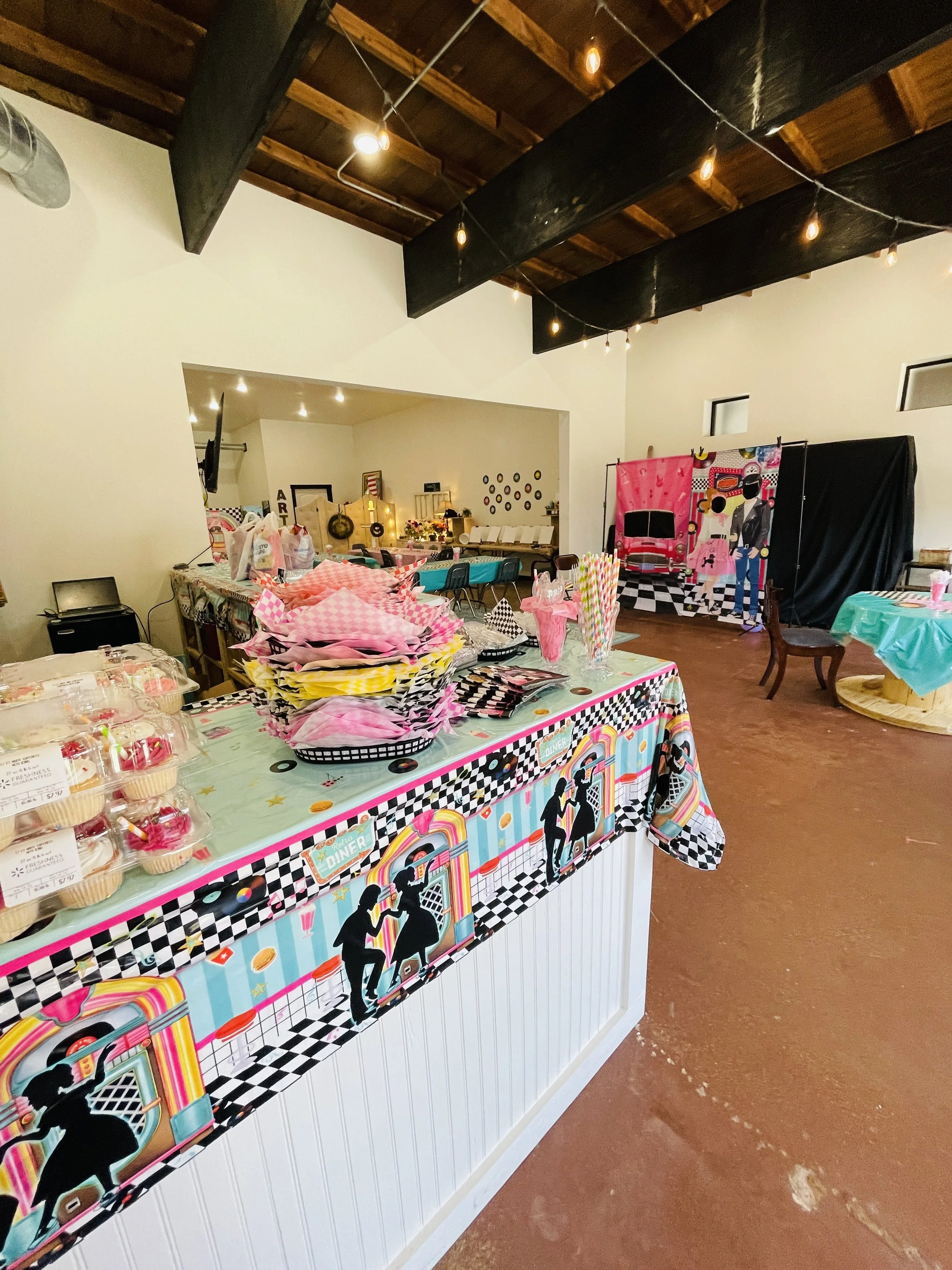 Decorated party setup with a colorful diner-themed tablecloth, pink and yellow napkins, and a photo backdrop featuring a retro diner scene.
