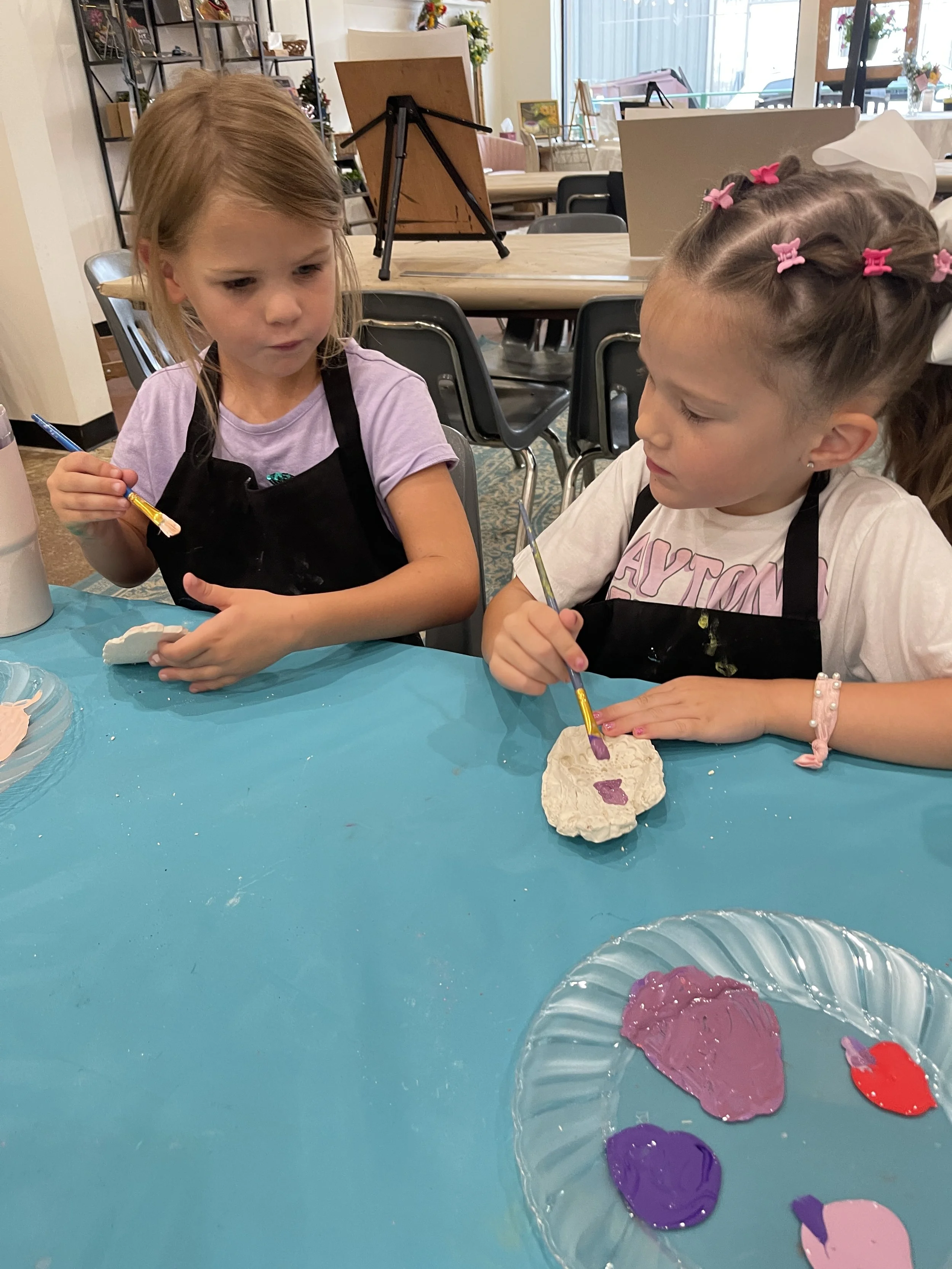 Two young girls are sitting at a table, painting rocks with purple and pink paint. They are focused on their craft projects, with one girl holding a paintbrush and the other looking at her painted rock. The table is covered with a turquoise tableclot