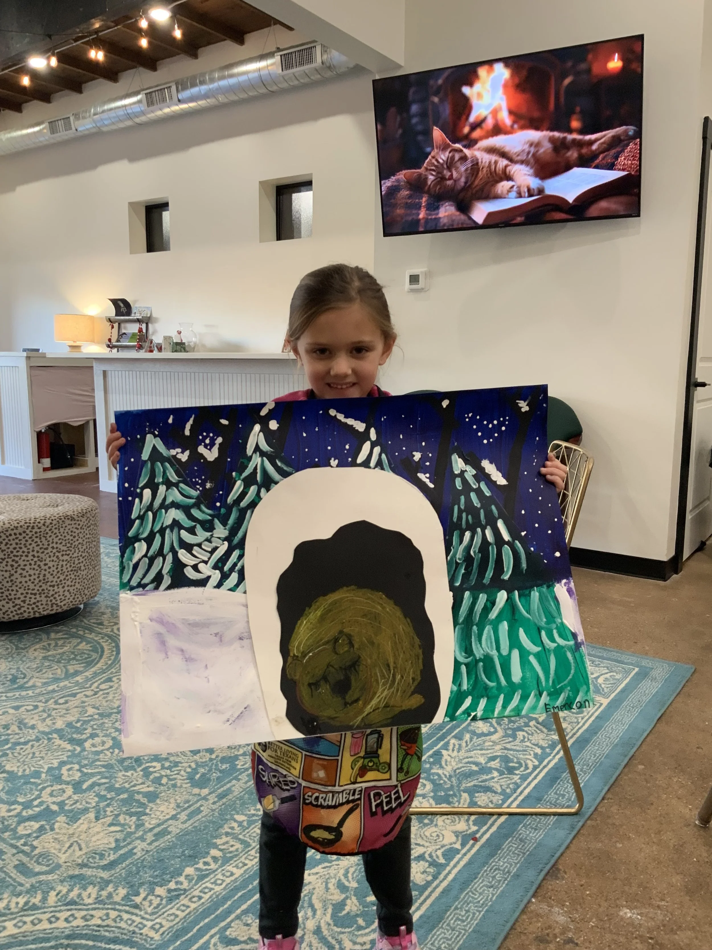 A young girl in pajamas holding a colorful painting of a bear in a forest at night, with a landscape including trees and a moonlit sky, inside a cozy room with a patterned rug, a TV on the wall, and ambient lighting.