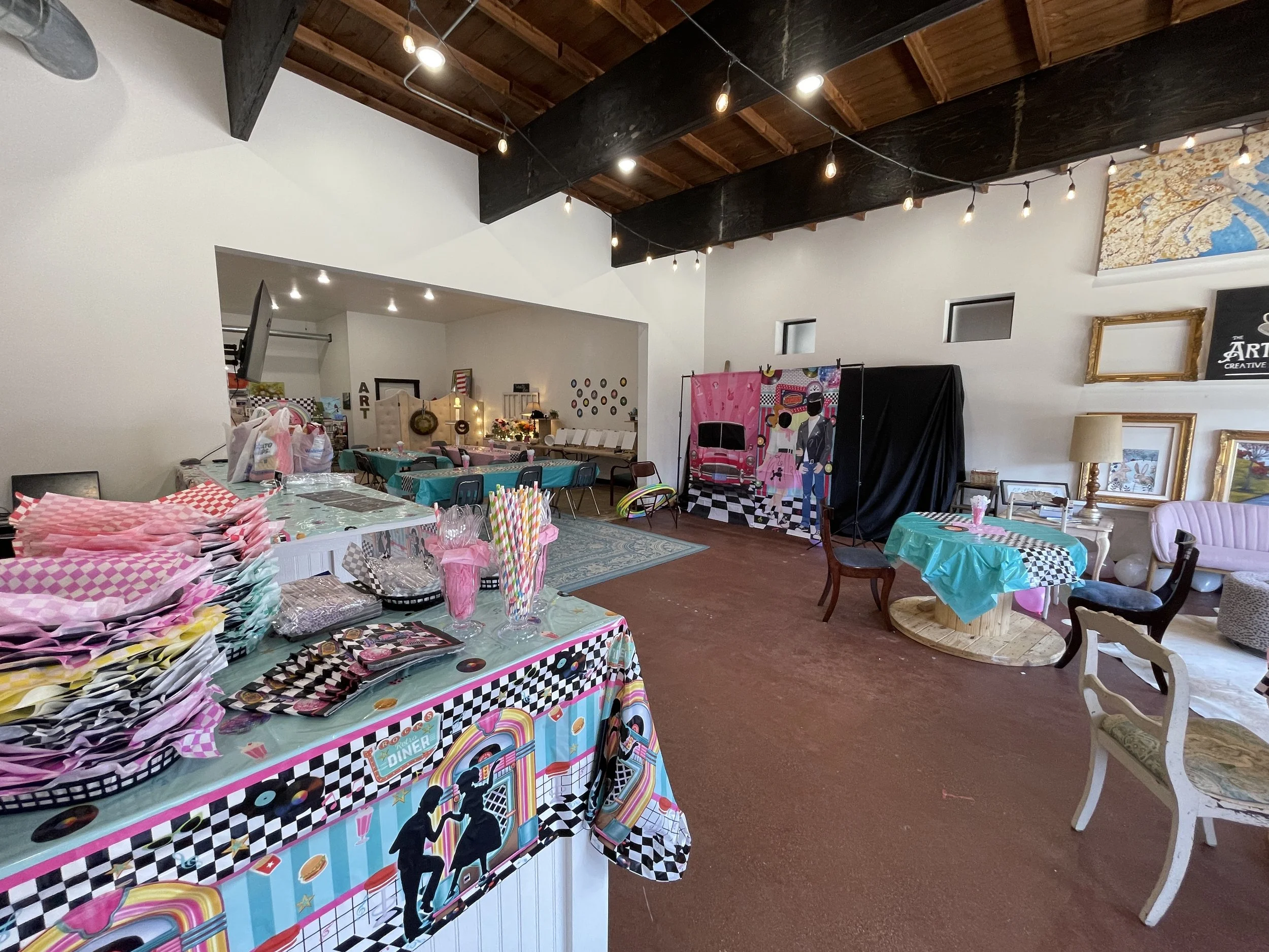 Colorful room decorated for a party with art, tables, and chairs, featuring pink and pastel themed decorations and a photobooth backdrop.