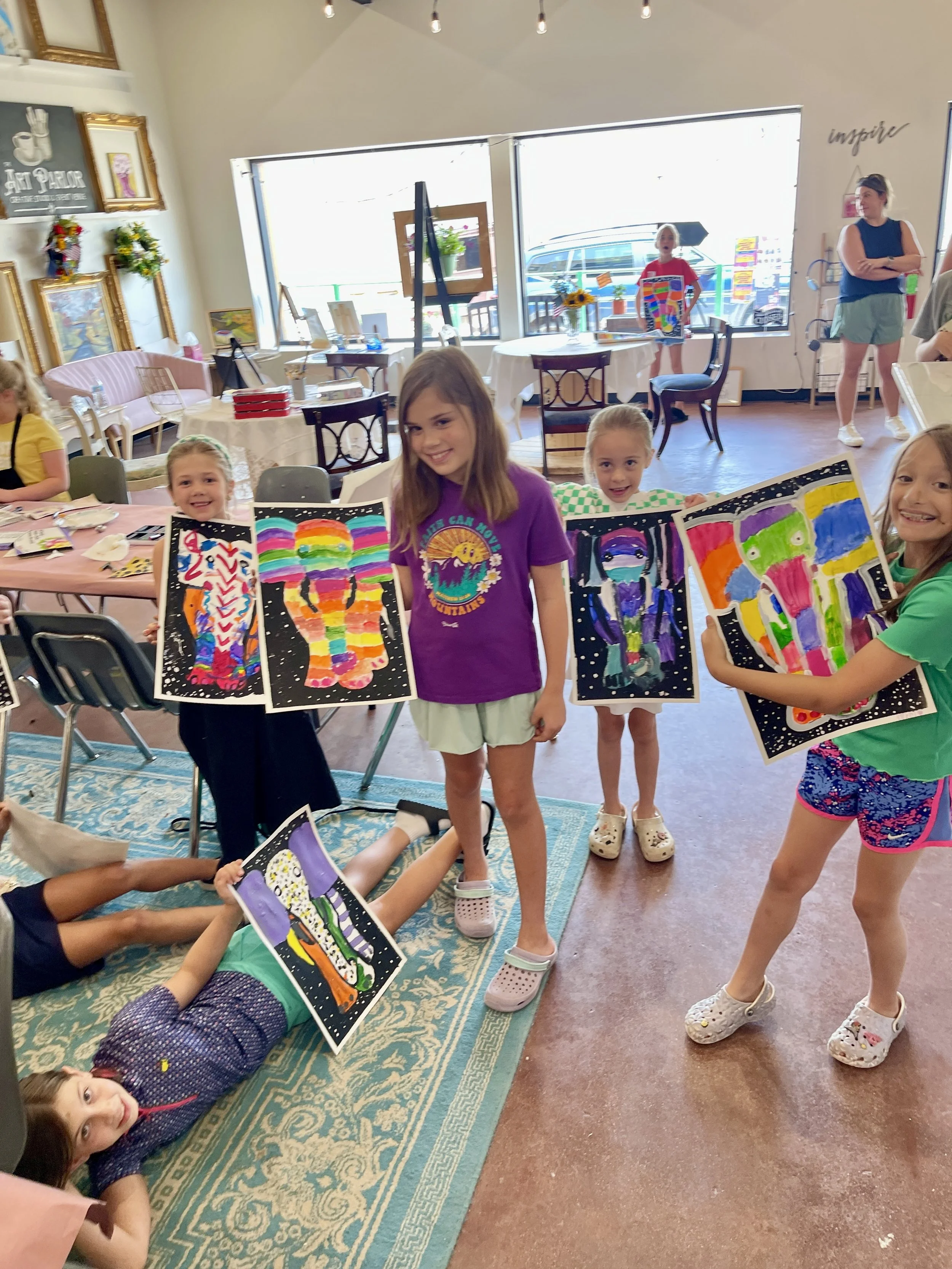 Children inside an art gallery holding colorful paintings of abstract and animal-like figures, with some children lying on a rug and others smiling at the camera.