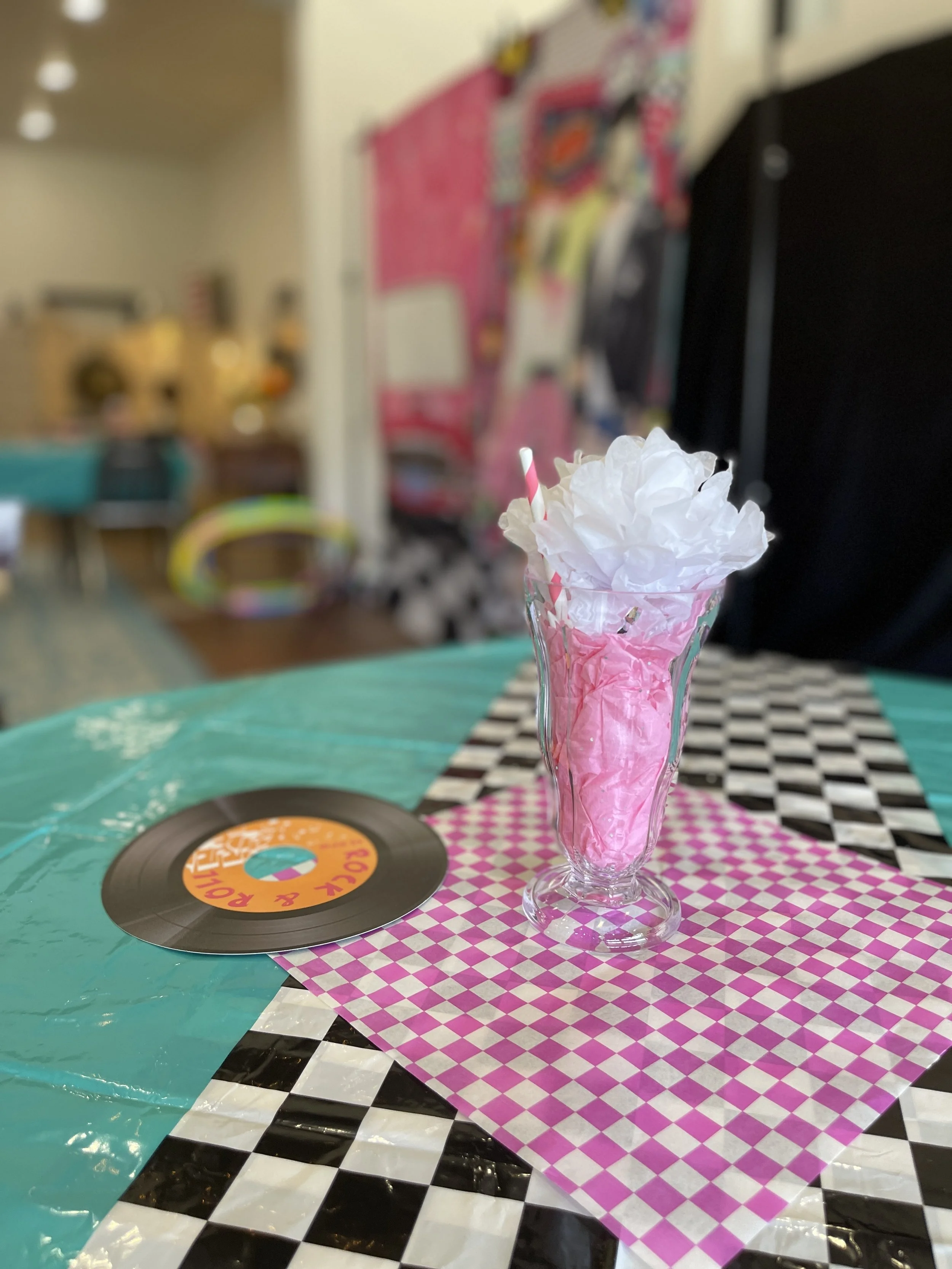 A tall glass with pink and white tissue paper, a pink and white striped straw, and white tissue paper on top, placed on a pink and white checkered napkin on a table with black and white checkered, teal, and black and white patterned tablecloths. A vi