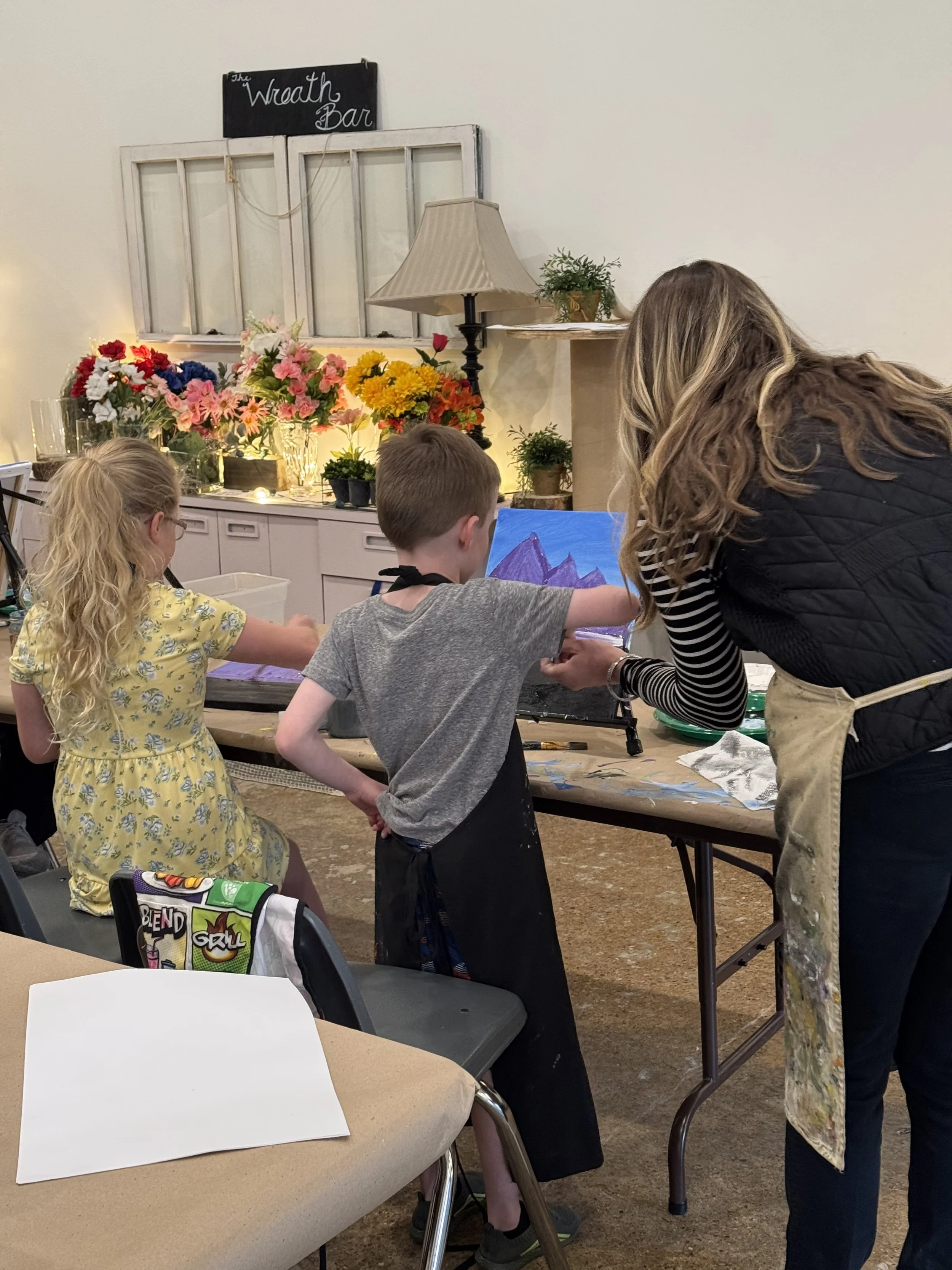 A woman assisting two children with painting at an art table, with colorful flowers and a lamp in the background. There's a sign that says 'The Wraith Bar' on the wall.