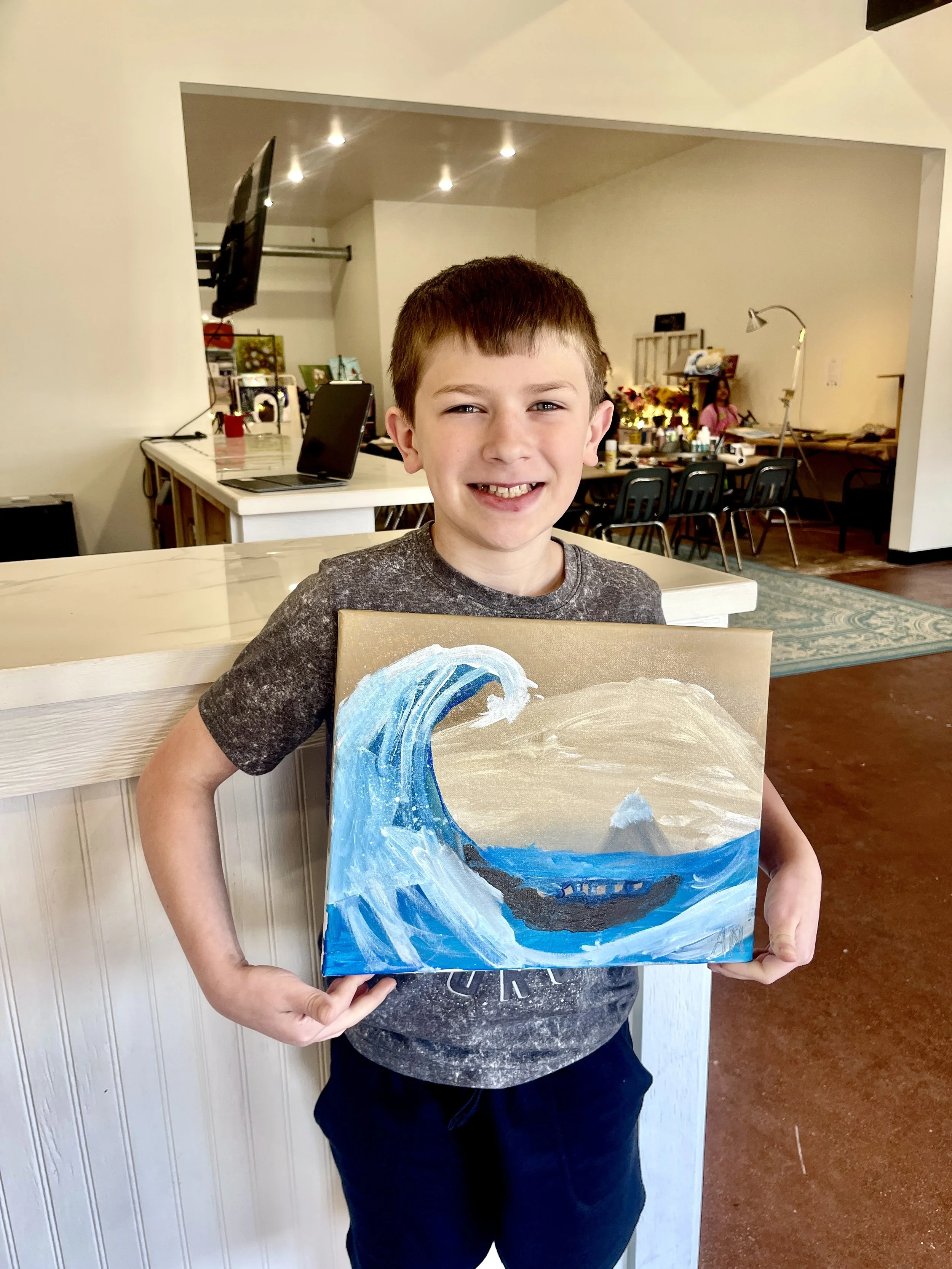 A smiling young boy holding a painting of a large ocean wave and a boat.