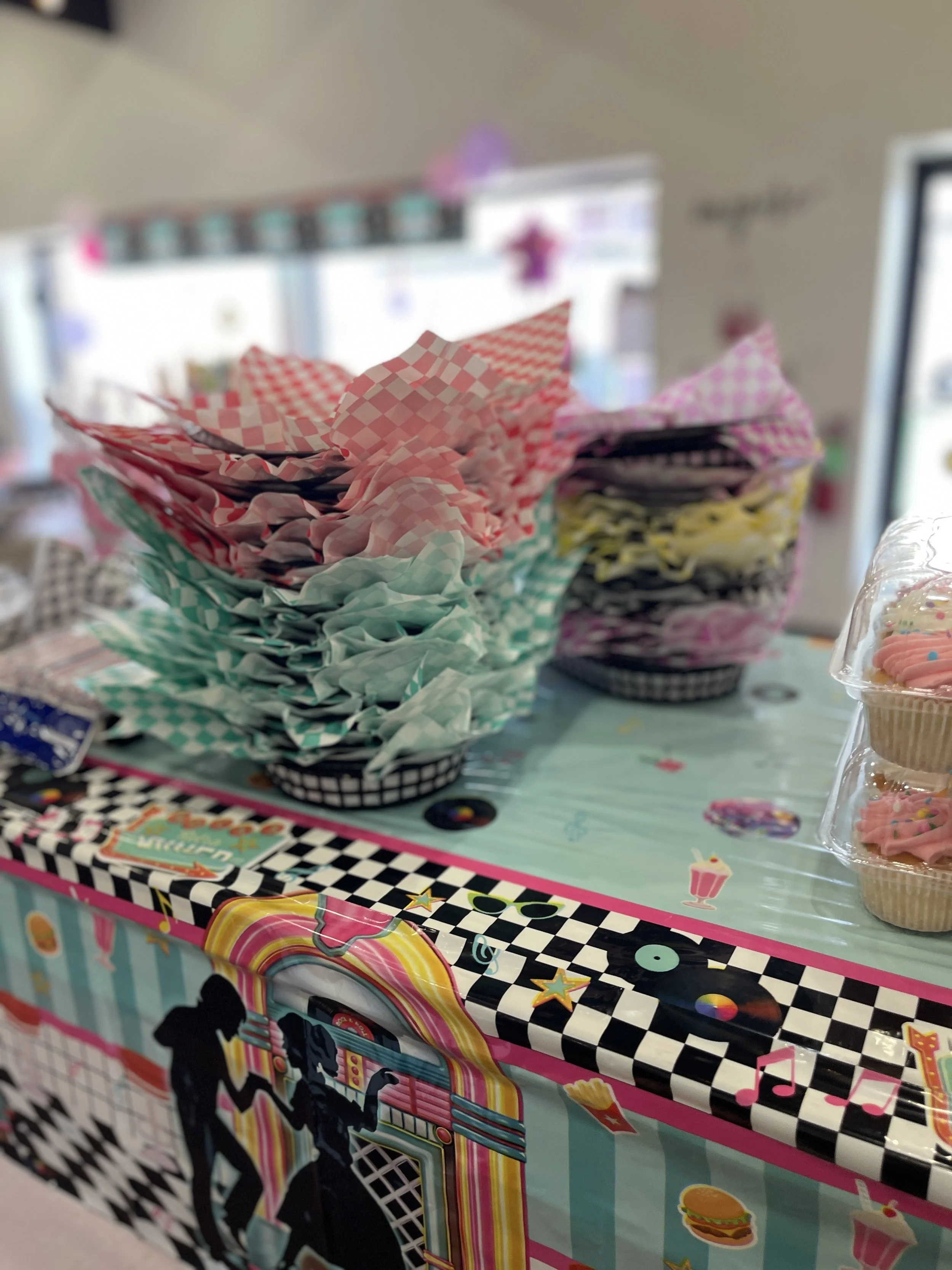 Colorful candy or snack bags stacked in checkered paper holders on a table decorated with a retro jukebox theme at a party or celebration.