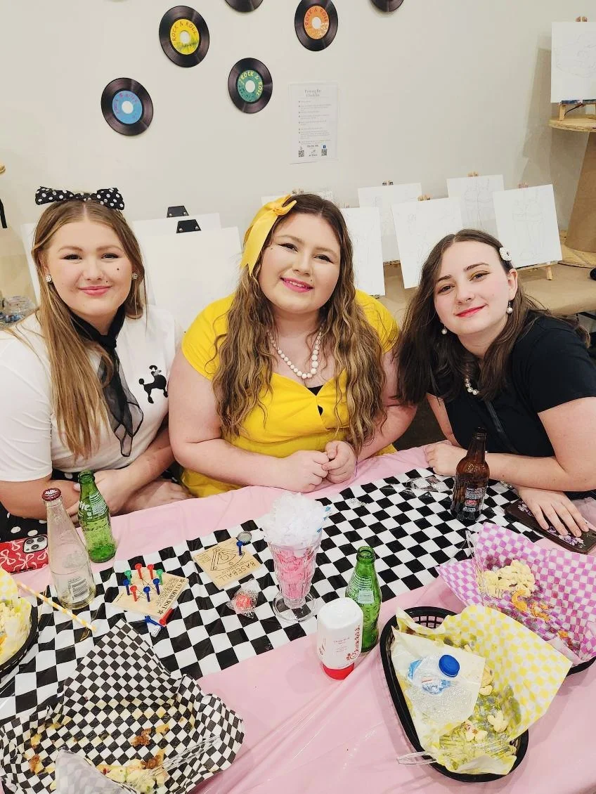 Three women sitting at a birthday party table with party food, drinks, and cakes. The table has a black and white checkered tablecloth, and there are pink decorations. The background features wall decorations of vinyl records and sketches on easels.