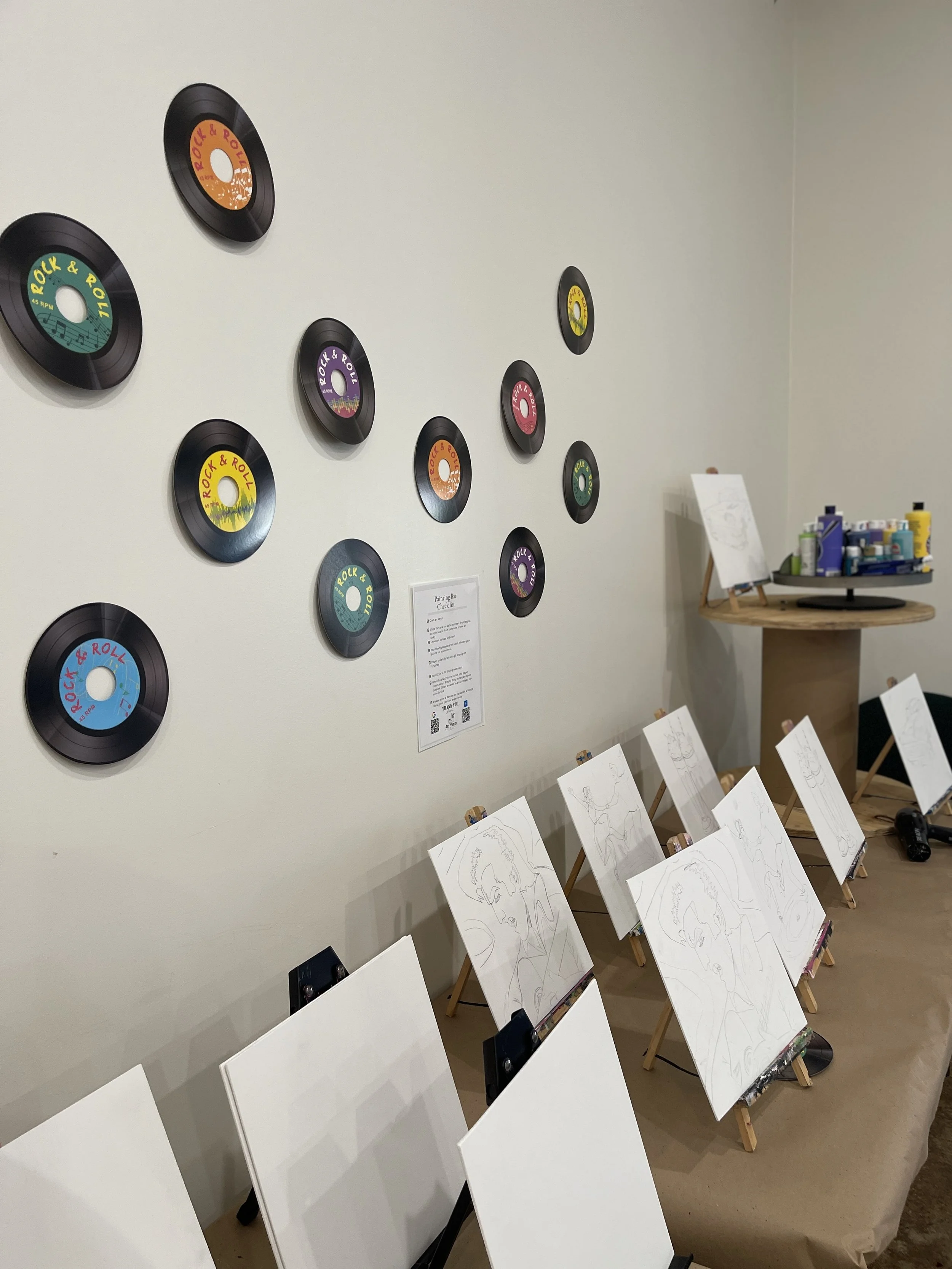 Room with multiple framed vinyl record wall decorations with colorful labels and several blank sketch canvases on easels, likely set up for a painting class or art event.