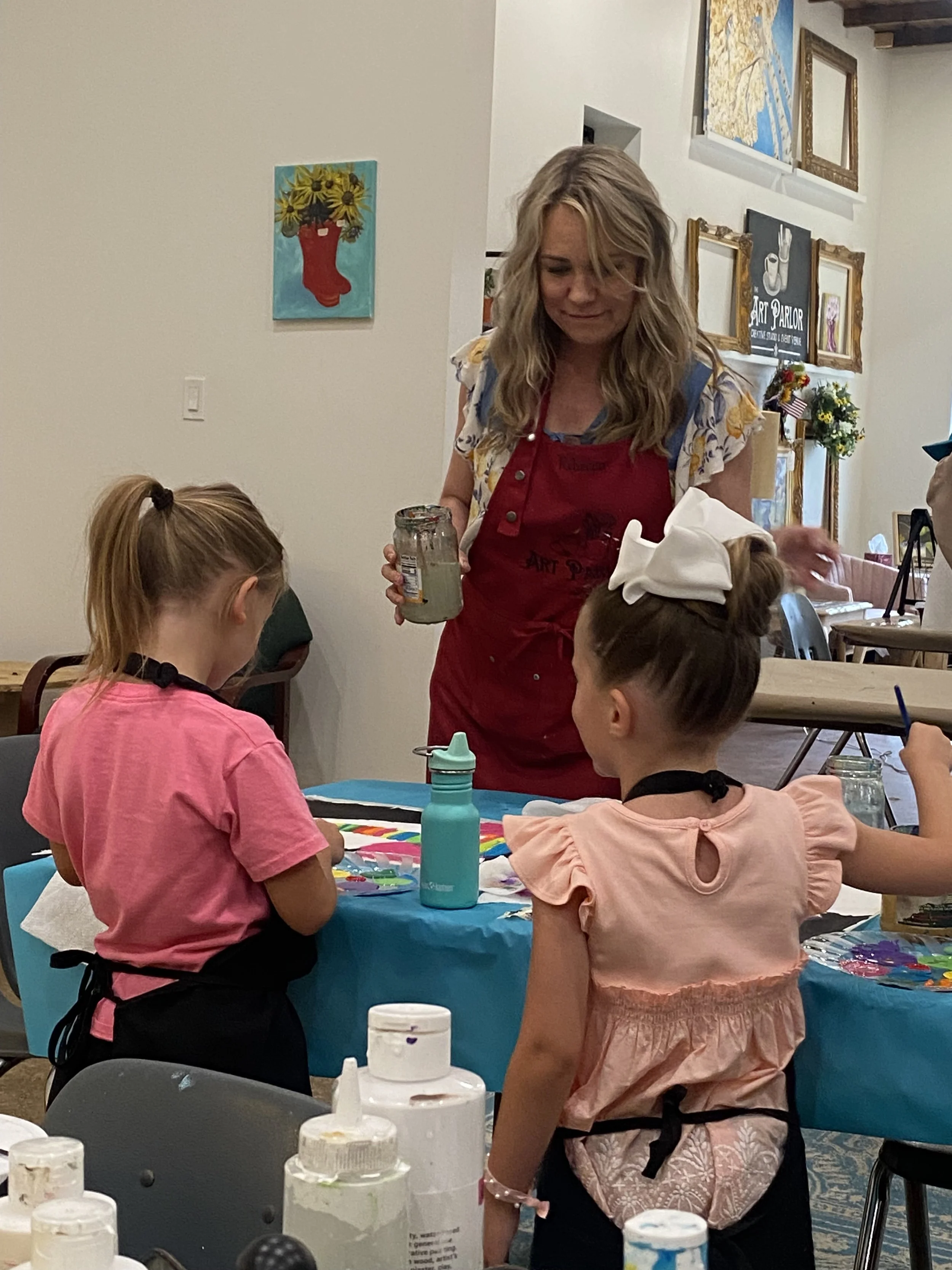 A woman in a red apron teaching acrylic painting to two young girls at a table in an art studio or classroom.