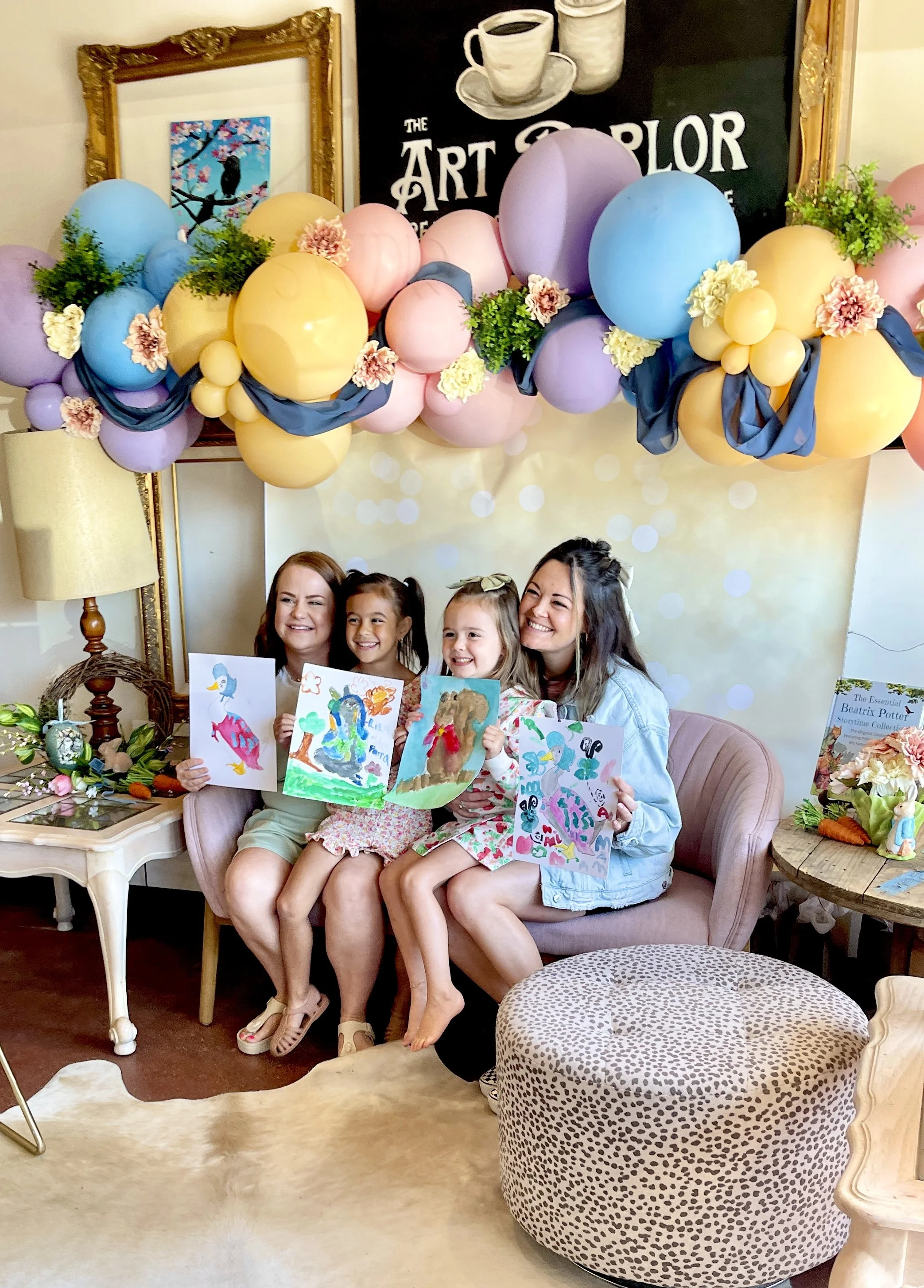 Four women and three young girls sitting on a pink sofa at a children's art party, holding colorful drawings of animals, with pastel balloons and decorations overhead.