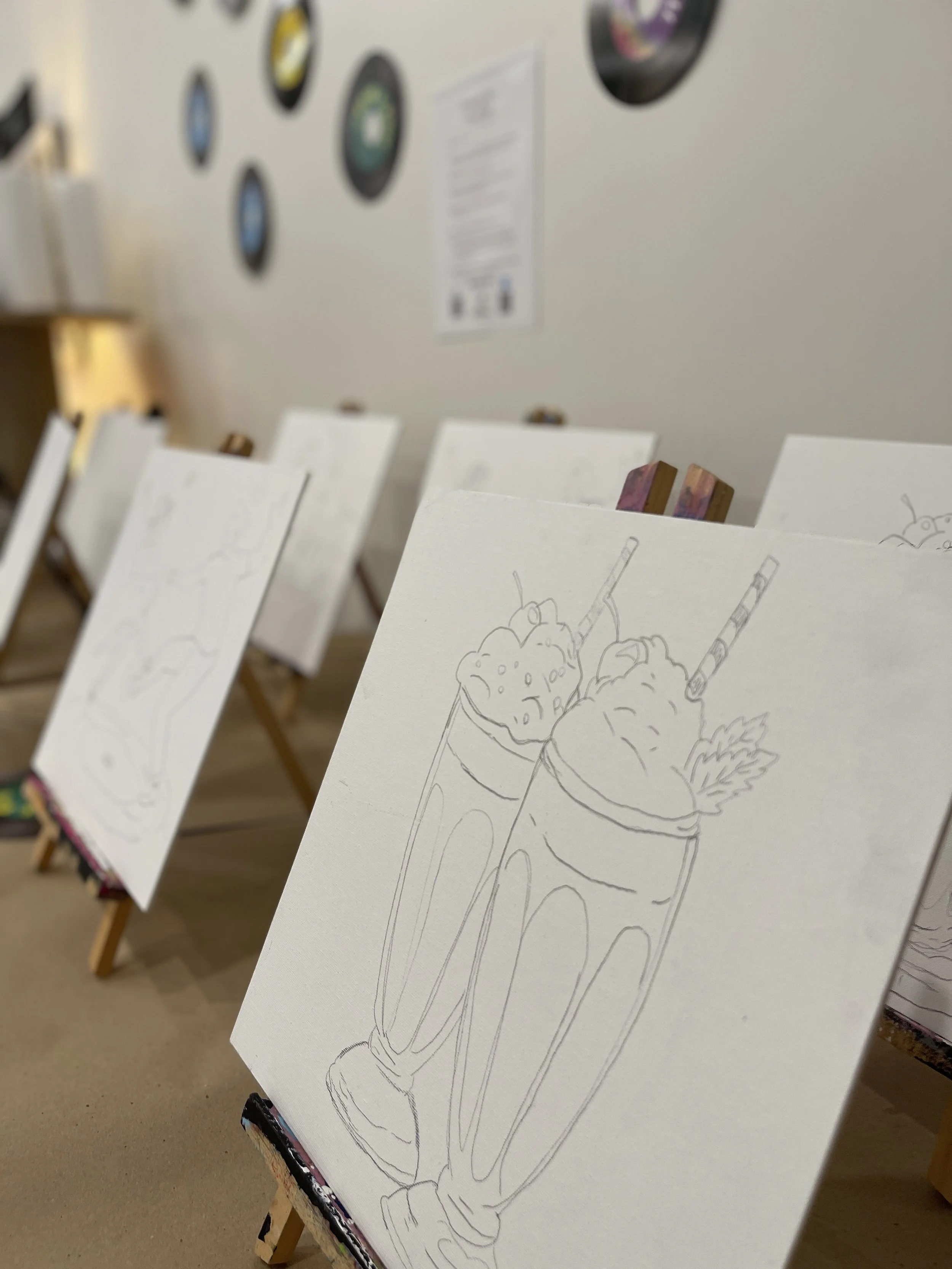 Sketch of two milkshakes with whipped cream, topped with a straw and a leaf, on a blank canvas in an art studio.