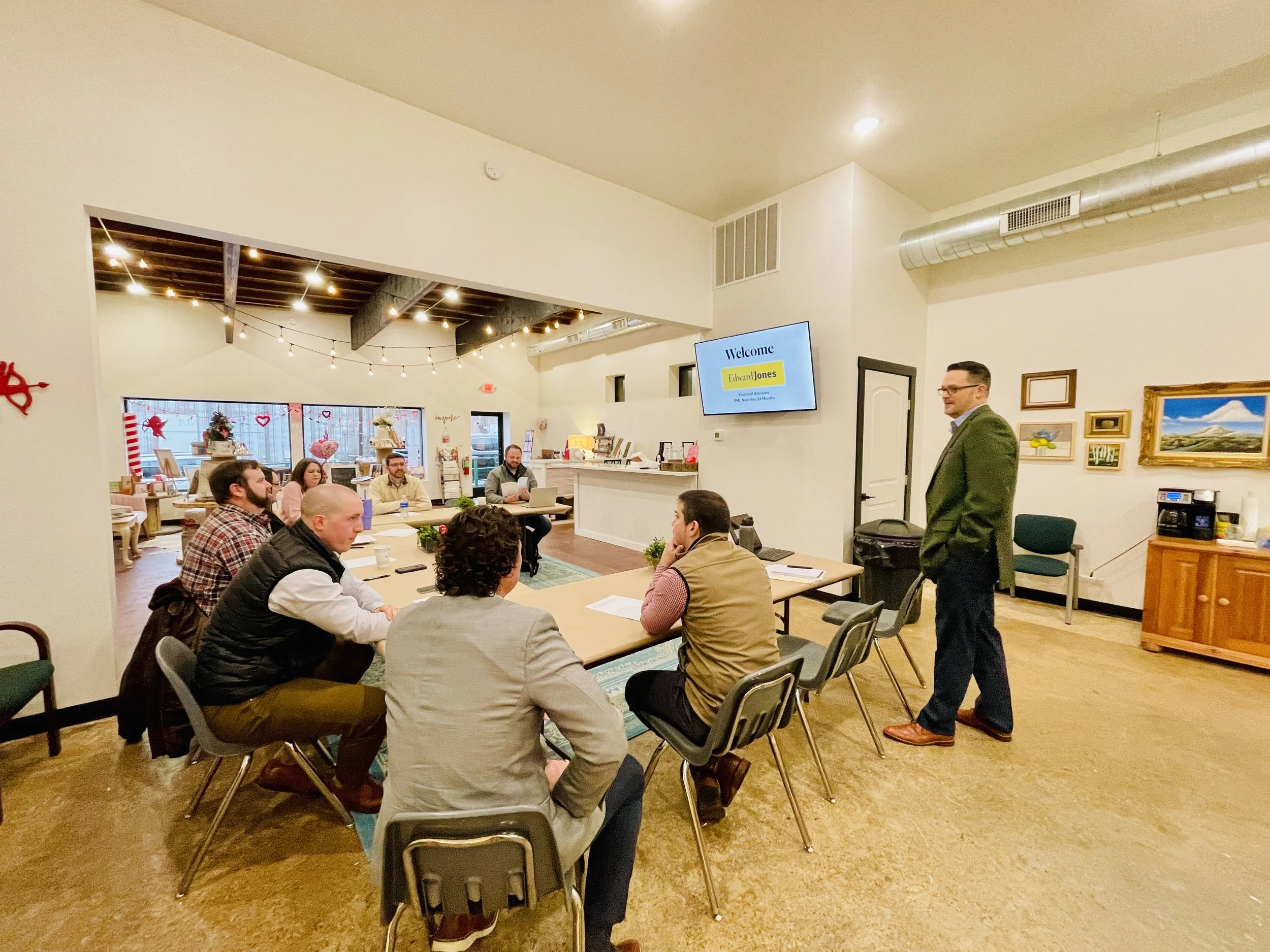 A group of people seated around a conference table in a meeting room, with a man standing near a TV screen that displays a welcome message, and a decorated area in the background with holiday decorations.