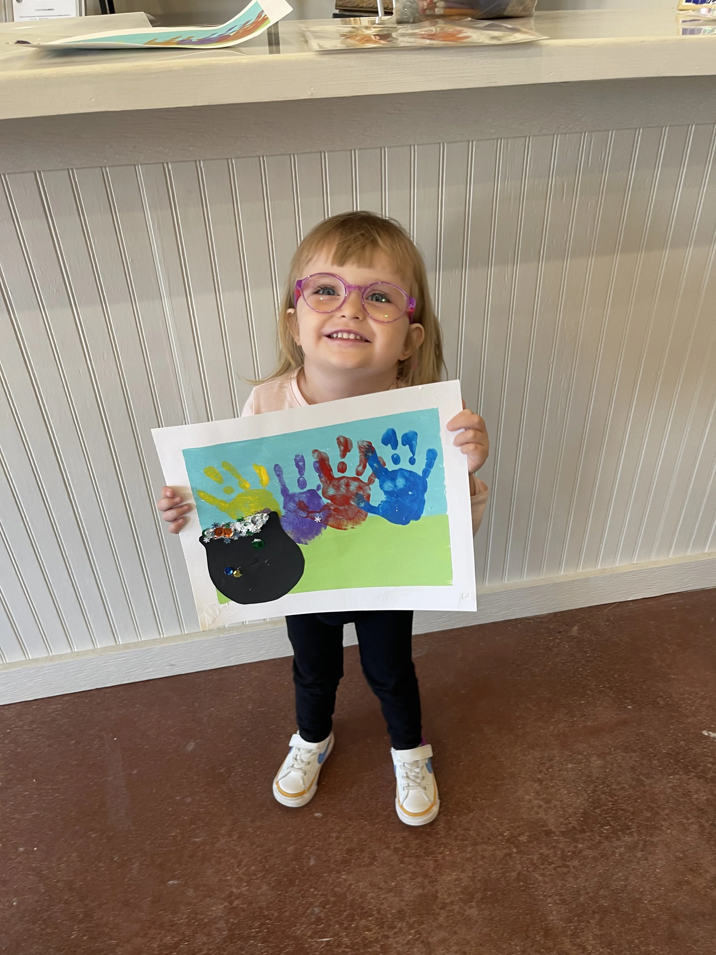 A young girl with glasses holding a colorful artwork featuring painted handprints and a black circle decorated with small objects. She is smiling and standing indoors with a white paneled wall and a counter behind her.