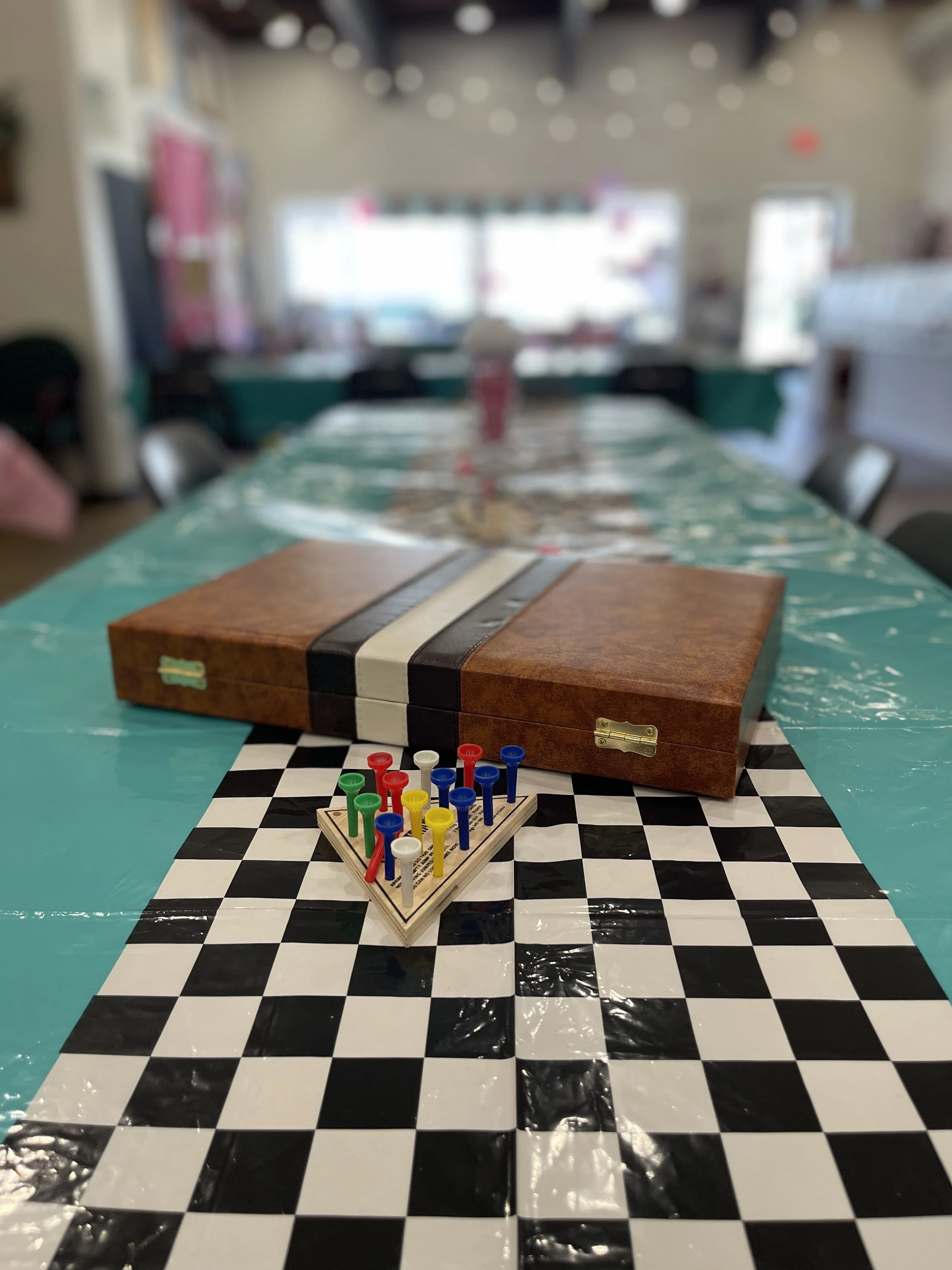 A game setup with a chessboard, a blue and white game piece holder, a brown wooden box, and a plastic tablecloth on a long table in a room with bright windows.