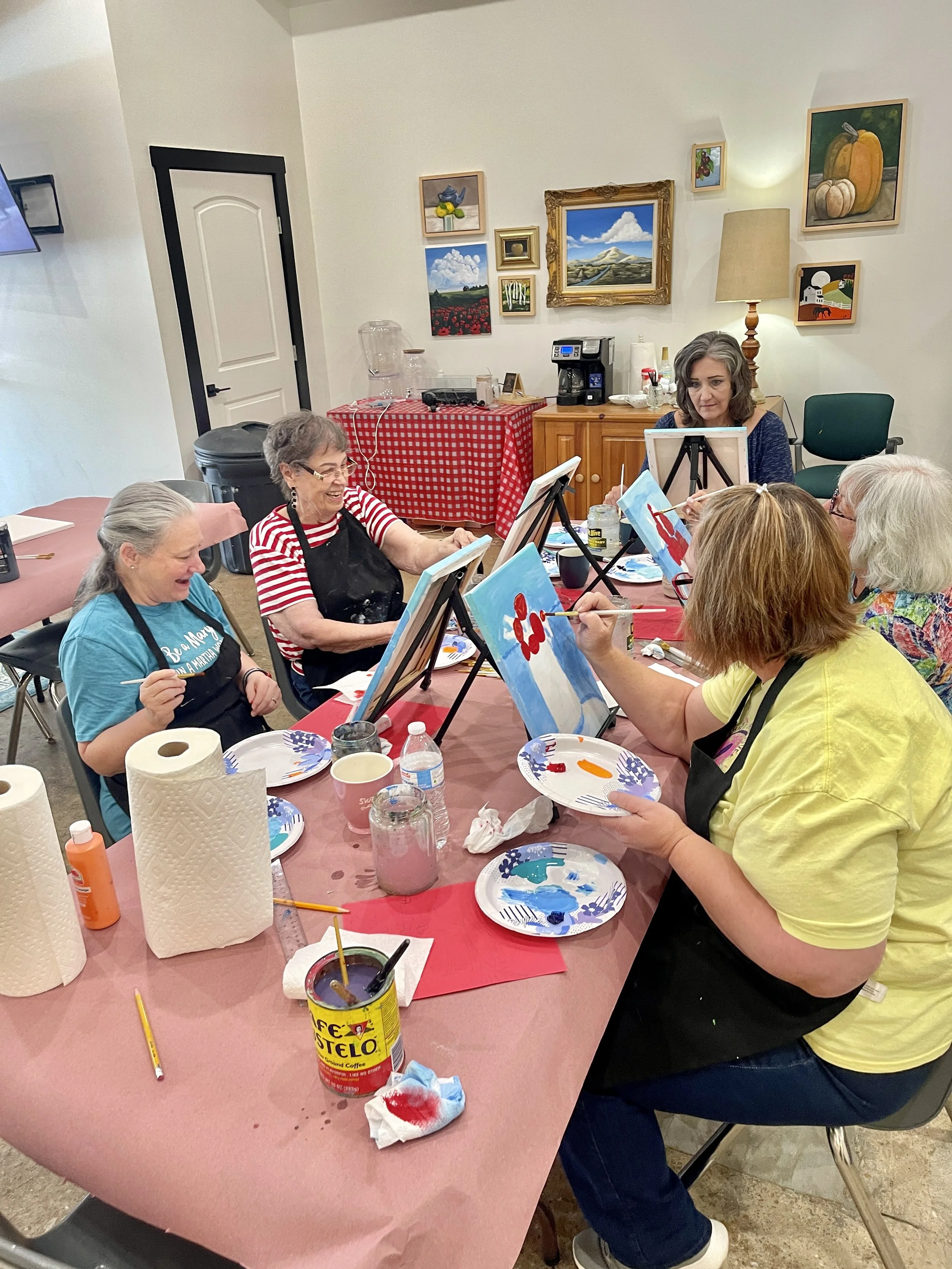 Group of women painting on canvases at a party or class, with painting supplies and beverages on the table.