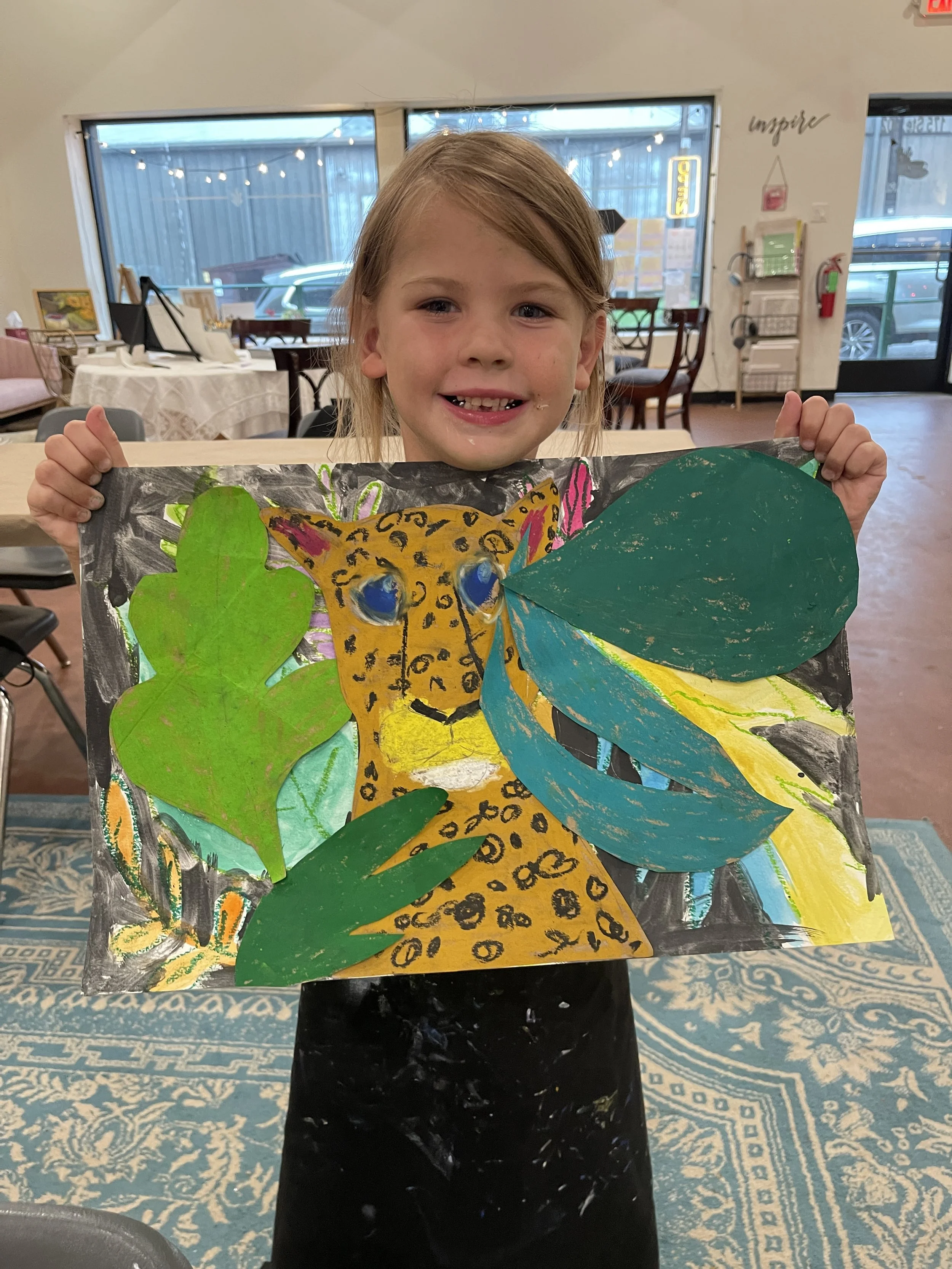 A young girl holding up a colorful drawing of a leopard with large green leaves and a blue and yellow background inside a restaurant or cafe.