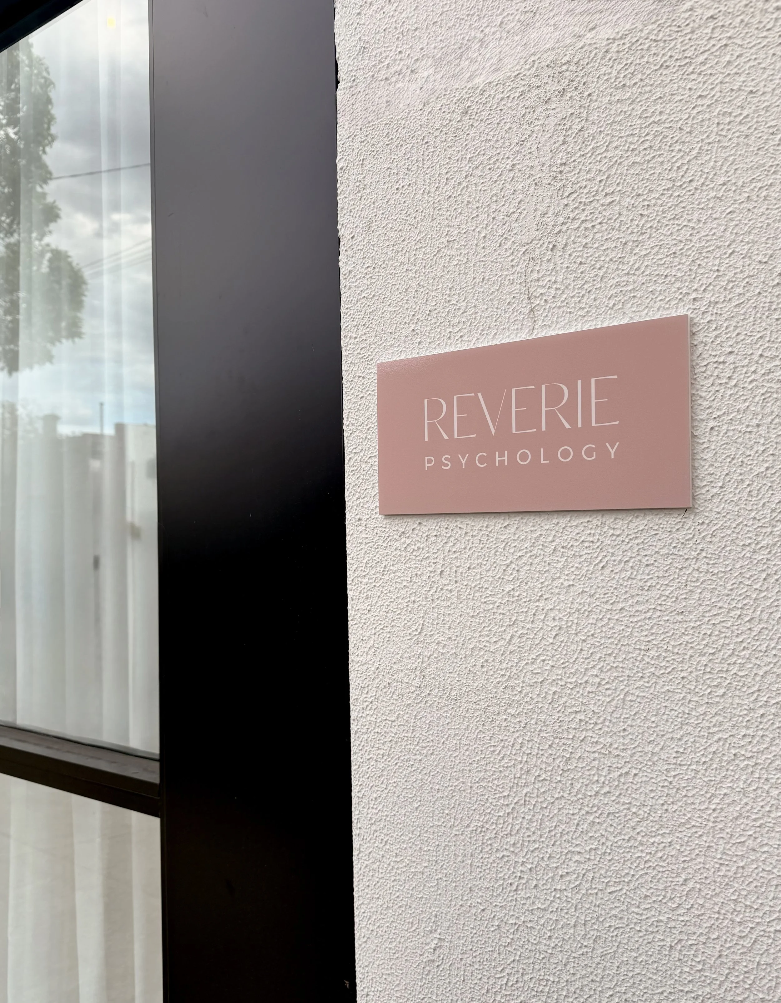 Close-up of a pink sign on a textured white wall that reads 'REVERIE PSYCHOLOGY' with a window or glass panel reflecting trees and sky on the left side.