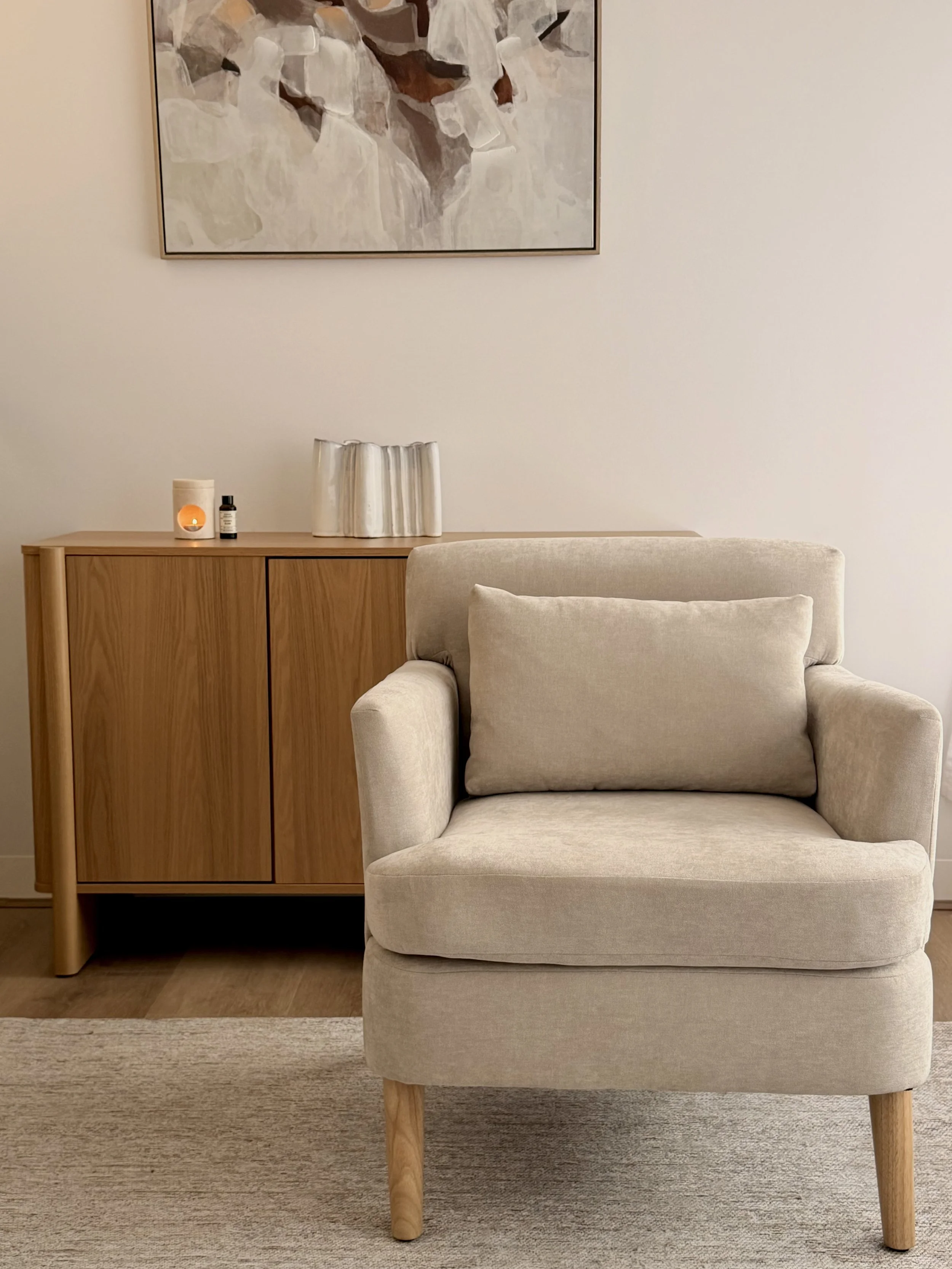 A beige upholstered armchair with wooden legs in front of a light-colored wall. A wooden sideboard with decorative items, including a white textured vase, small bottles, and a candle, is in the background. A framed abstract painting hangs on the wall above the sideboard.