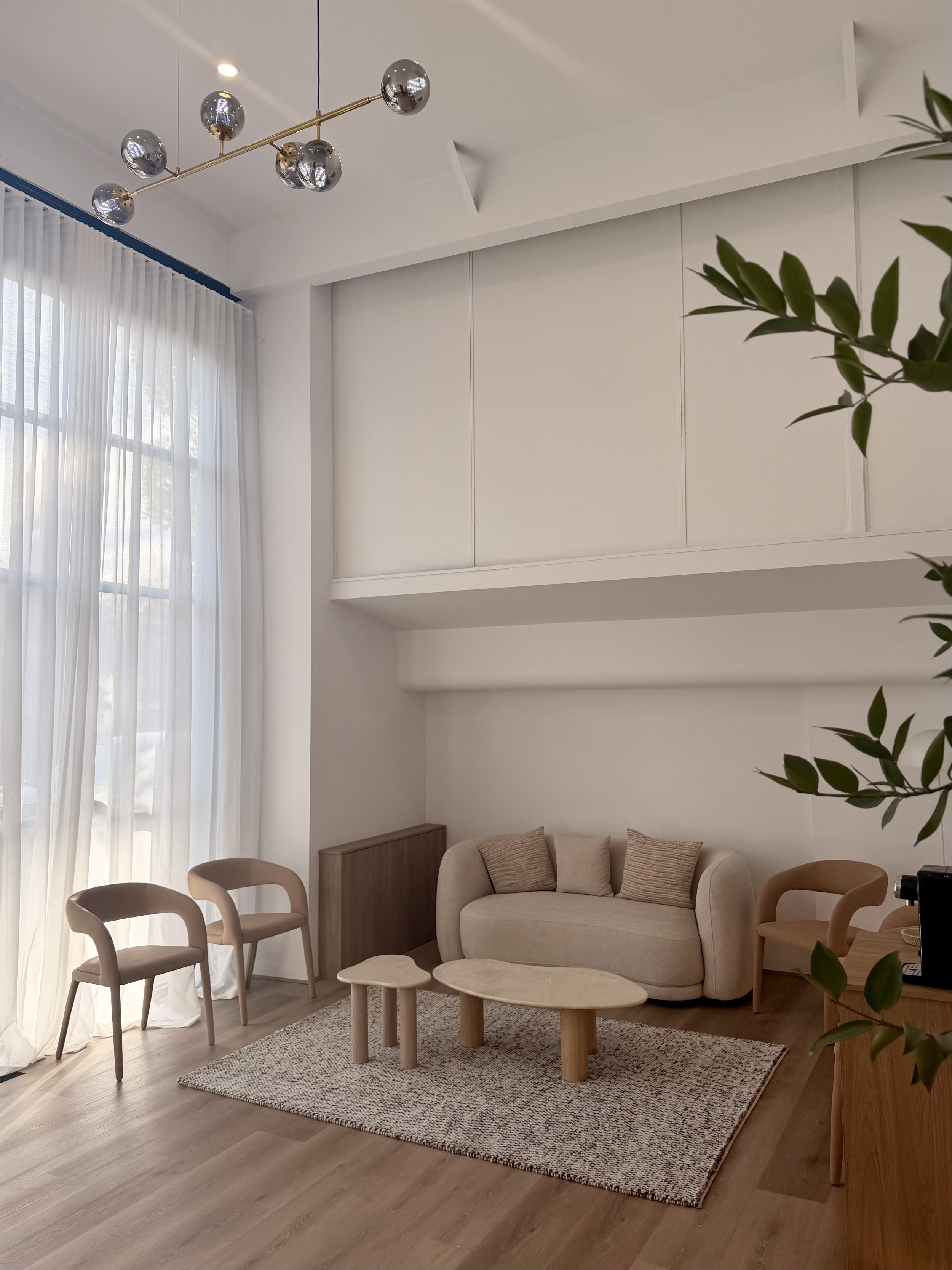 Modern living room with beige furniture, wooden chairs, small ottomans, a rug, large windows with white curtains, and a white wall with a built-in cabinet. Ceiling light fixture is metallic with multiple spherical bulbs.