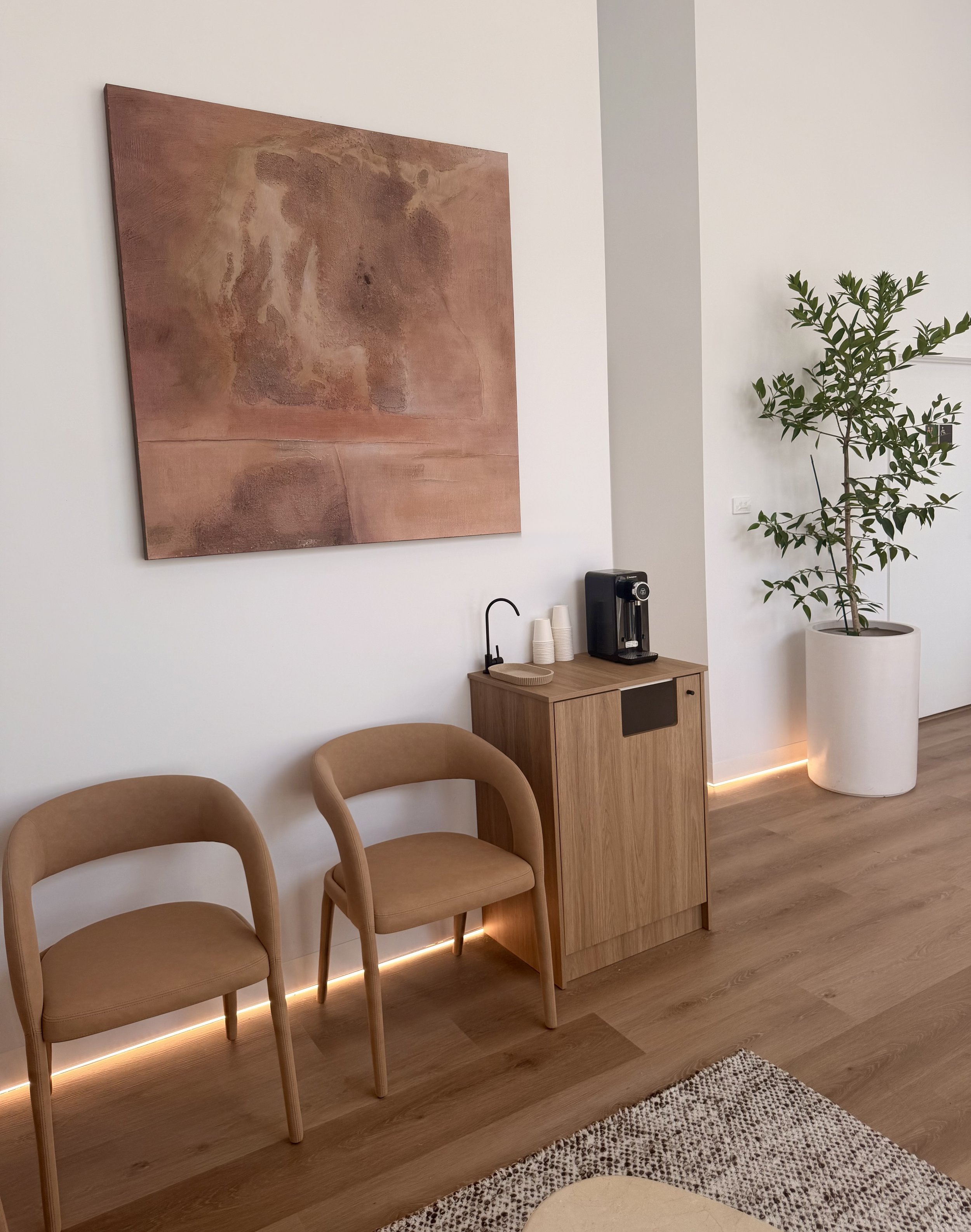 Interior space with beige chairs, a wooden cabinet with a coffee machine, cups and a sink, a large framed artwork on the wall, a potted plant, a patterned rug on the floor, and string lights beneath the furniture.