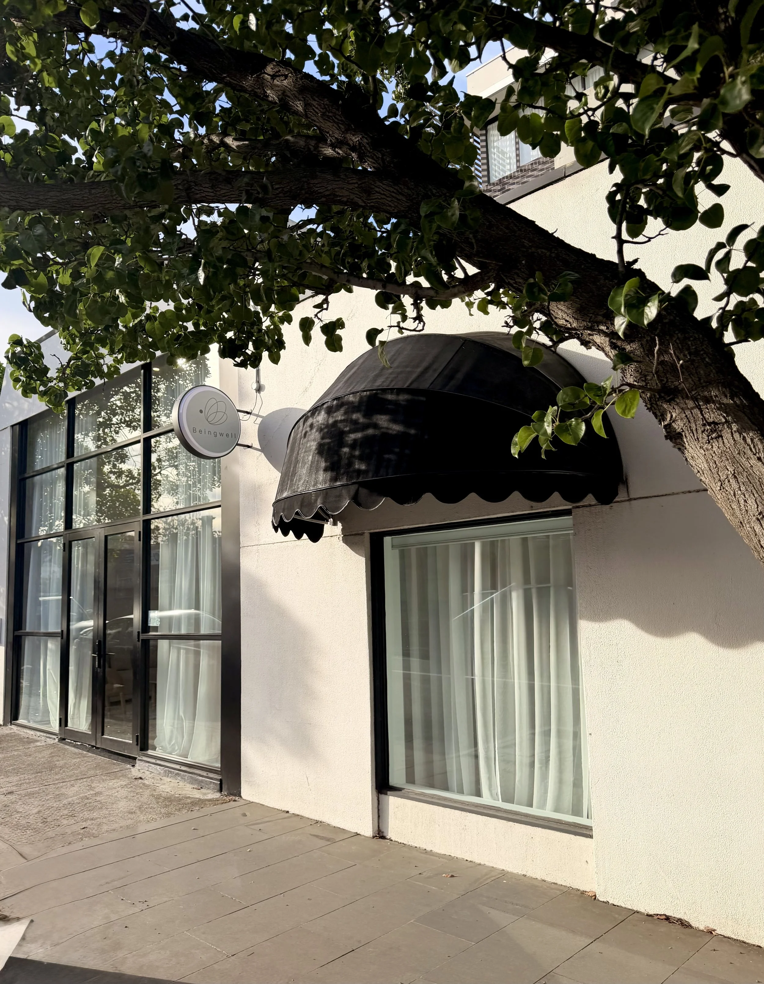 View of a building's exterior with large glass windows, a black awning, and a tree with broad leaves partially obscuring the view. The building has a sign that reads 'Beingwell'.