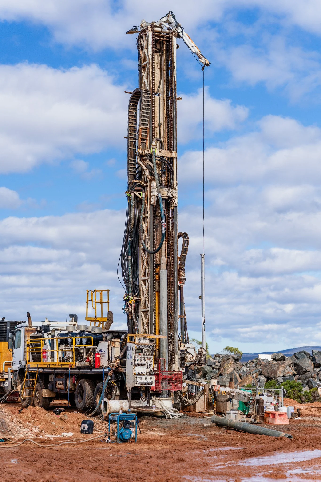 A large WA Water Bores drilling rig on a dirt site with rocks and muddy patches, with cloudy sky in the background.