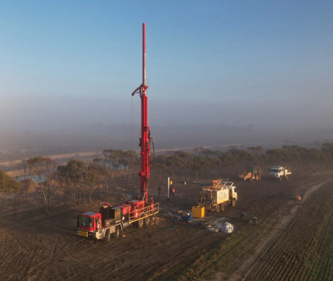 A large red and white drilling rig set up in a field with trees, surrounded by several trucks and equipment. The sky is hazy and the ground appears arid or recently disturbed.