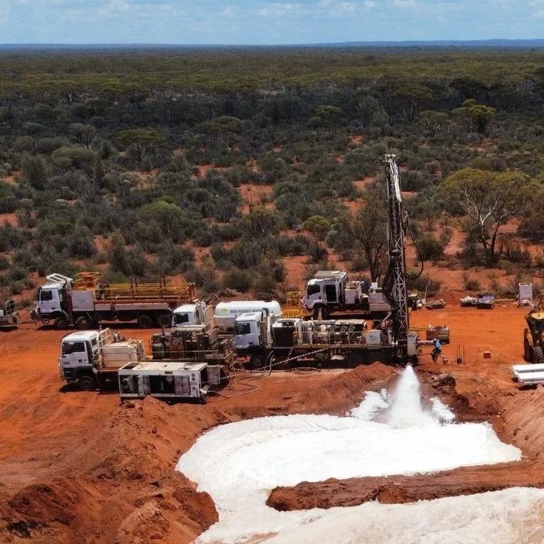 Drilling rig and support trucks on red dirt land with sparse trees, extracting liquid that is overflowing into a large pool.