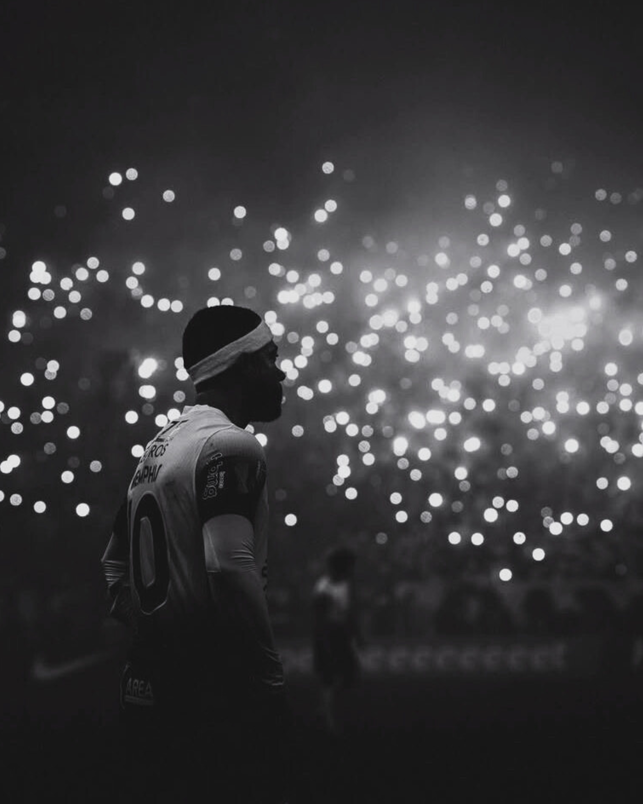 A silhouette of a soccer player with a white headband stands in front of a background of blurred lights, creating a bokeh effect, during a nighttime match.