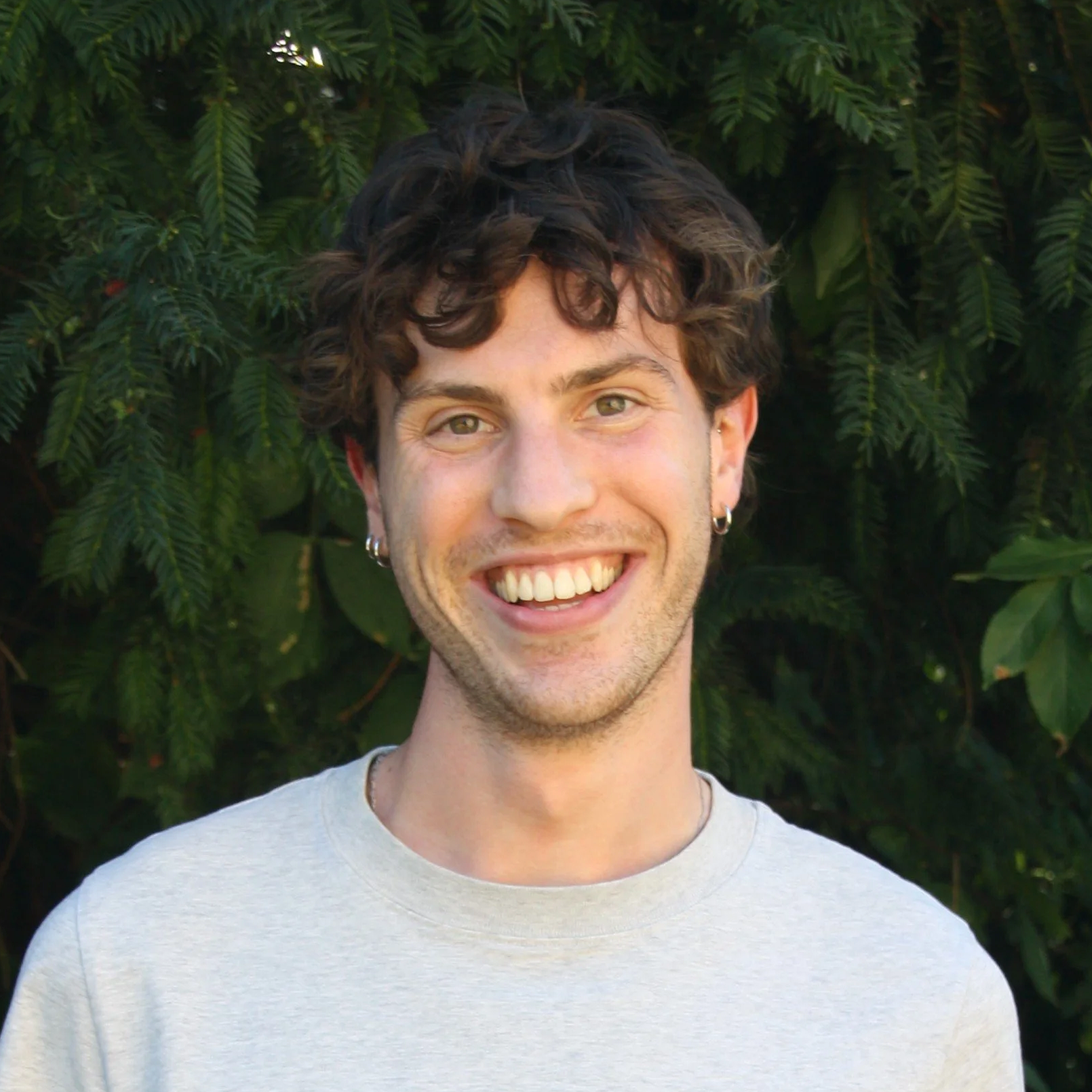 A young smiling man with dark curly hair standing outdoors in front of green leafy background.