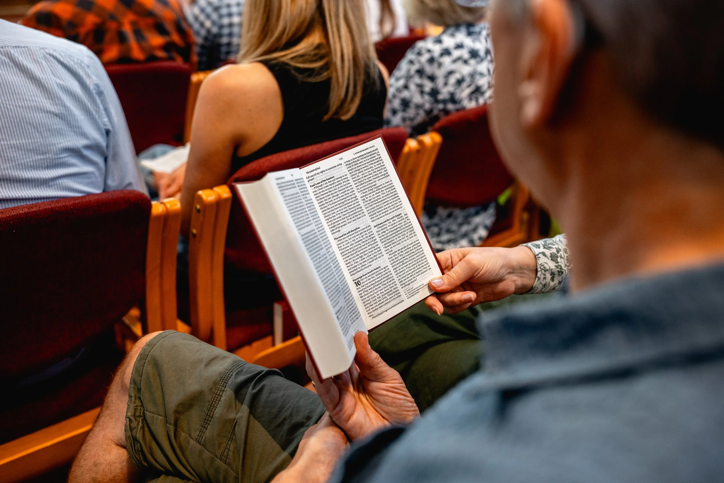 A person sitting in an auditorium reading a Bible while others sit nearby.