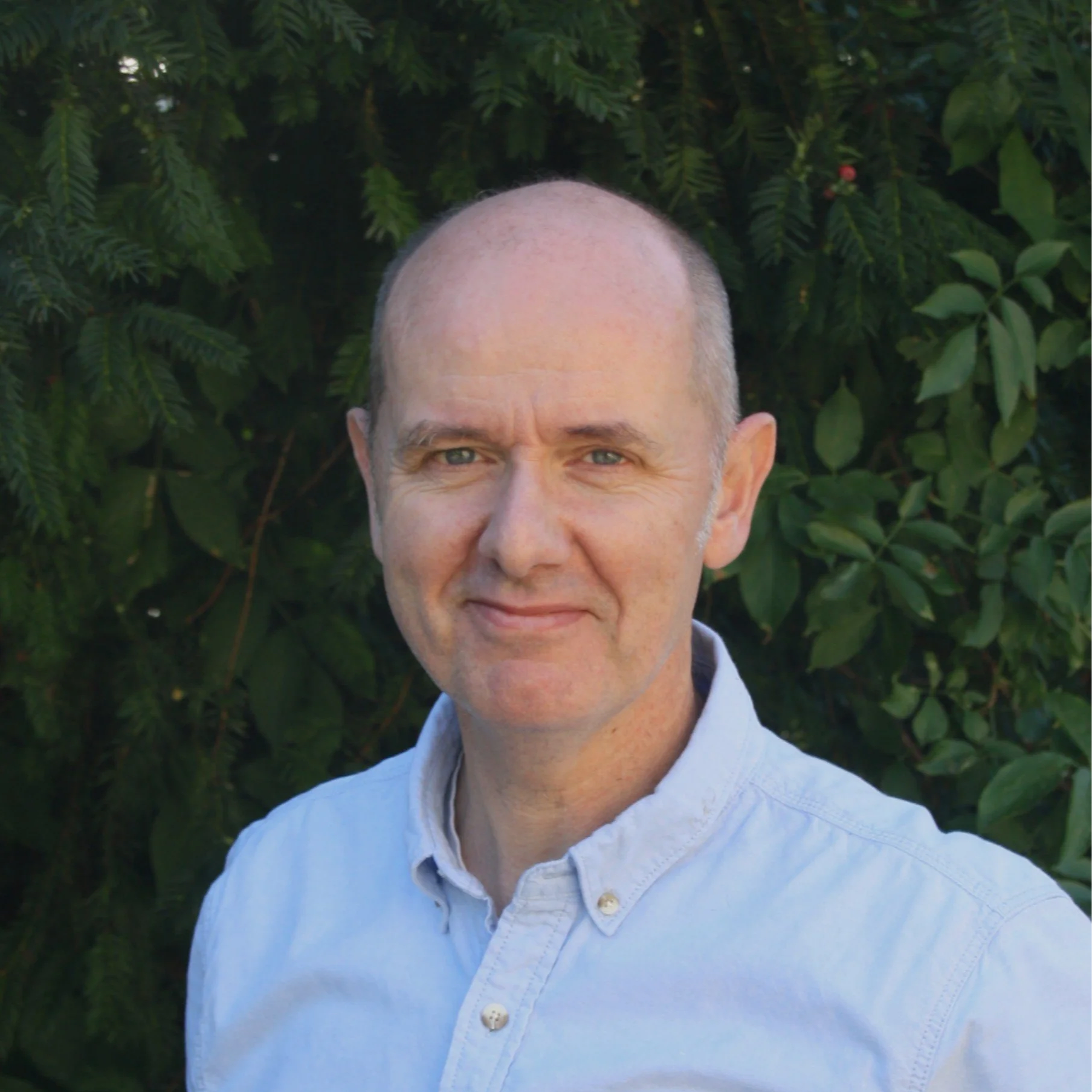 A man wearing a light blue button-up shirt, smiling slightly, standing outdoors in front of green foliage.