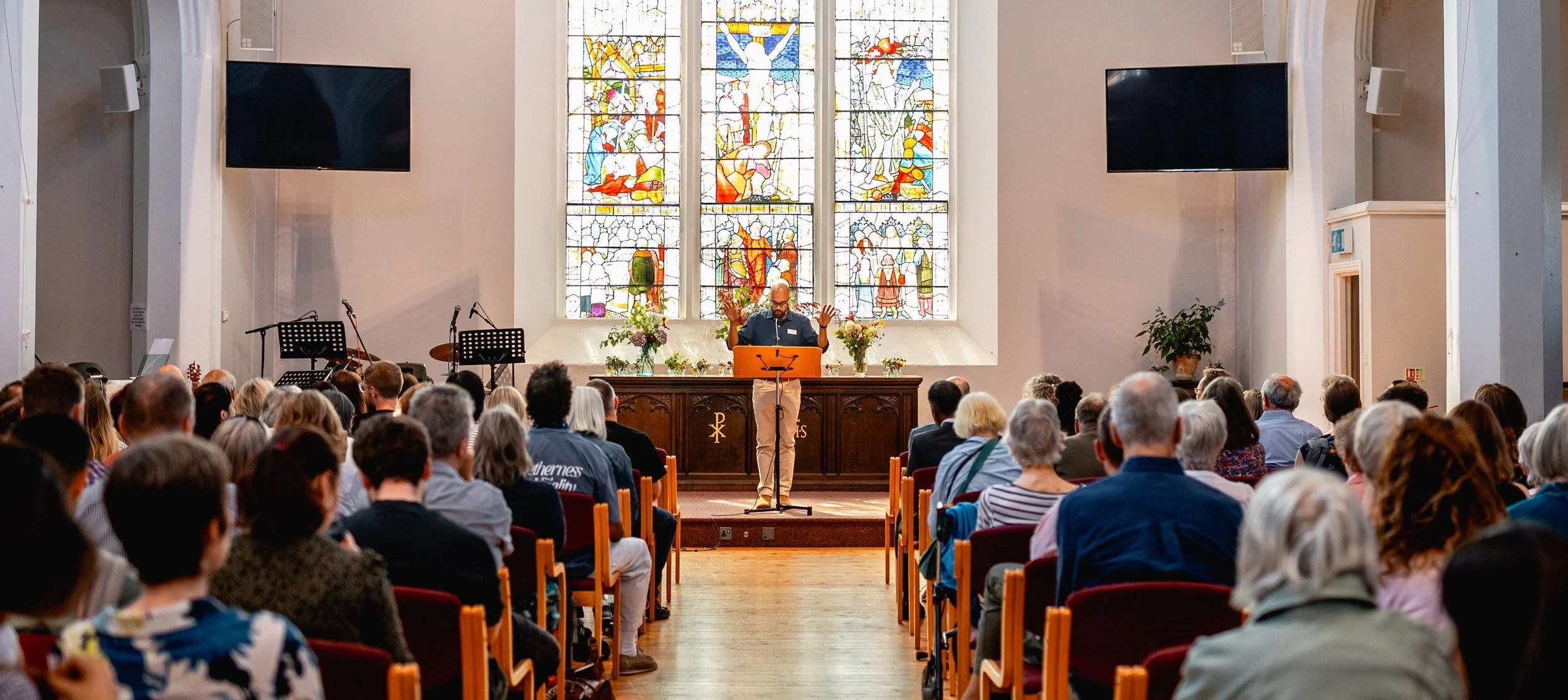 A preacher standing at a lectern inside a church with large stained glass windows behind him. The congregation is seated facing him, attentively listening. The church interior includes musical instruments and plants.