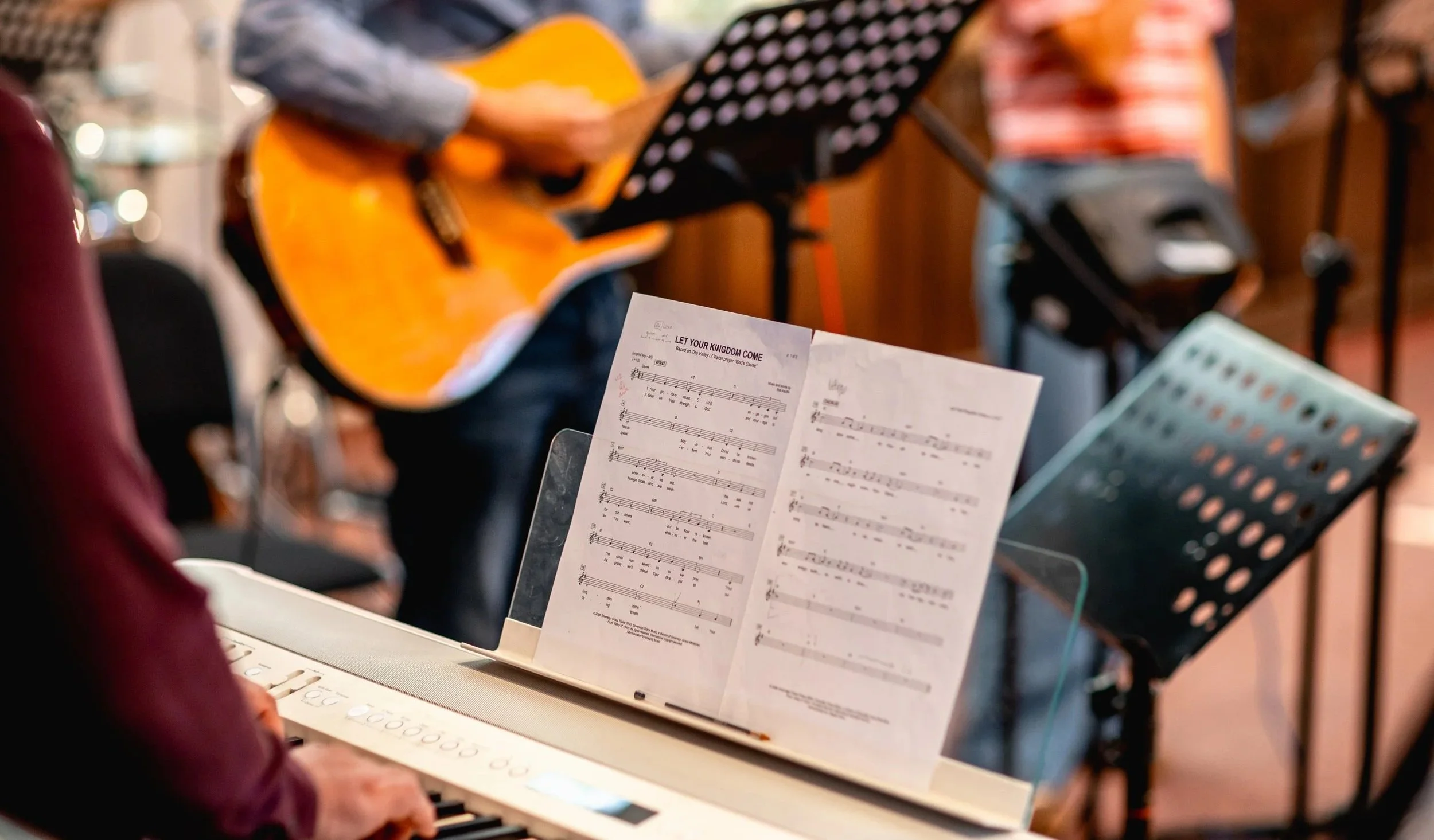 Close-up of a music stand with sheet music titled "Let Your Kingdom Come" in a group music performance setting, with a guitar and music stands visible in the blurred background.