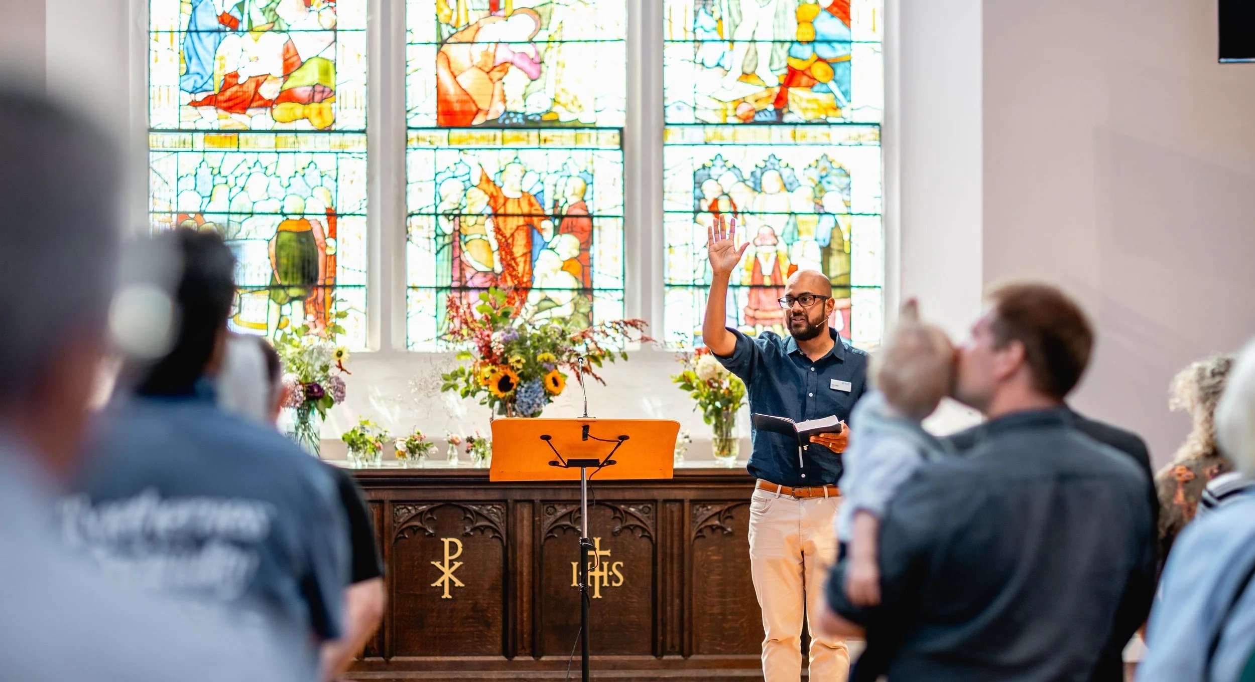 A man in glasses and a dark shirt is speaking and raising his hand inside a church with stained glass windows and floral arrangements.