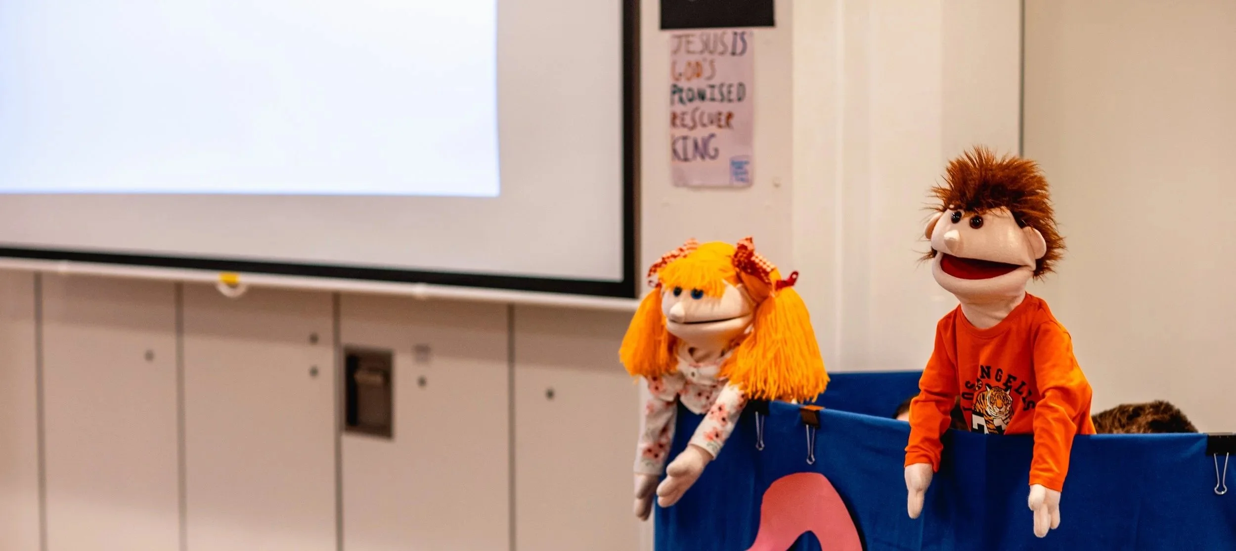 Two puppets with exaggerated facial features, orange hair, and dressed in orange shirts, sitting behind a blue puppet stage.