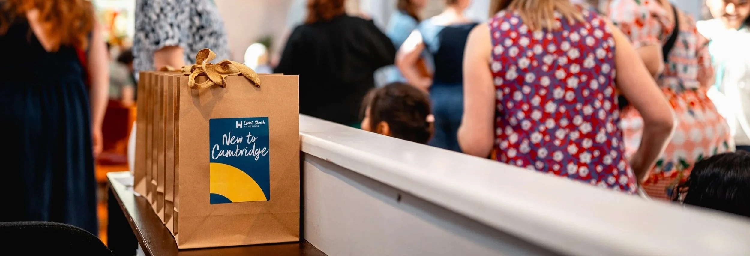 Brown paper gift bags with a label that says 'New to Cambridge' sitting on a table at a crowded indoor event.