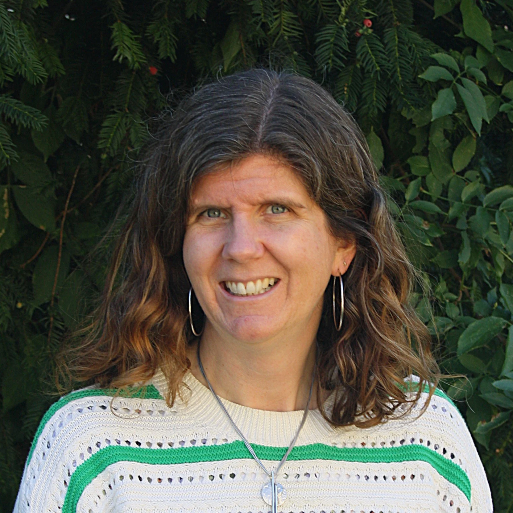 A woman with curly brown hair and blue eyes, wearing a cream-colored sweater with green and white stripes and earrings, smiling in front of green foliage.