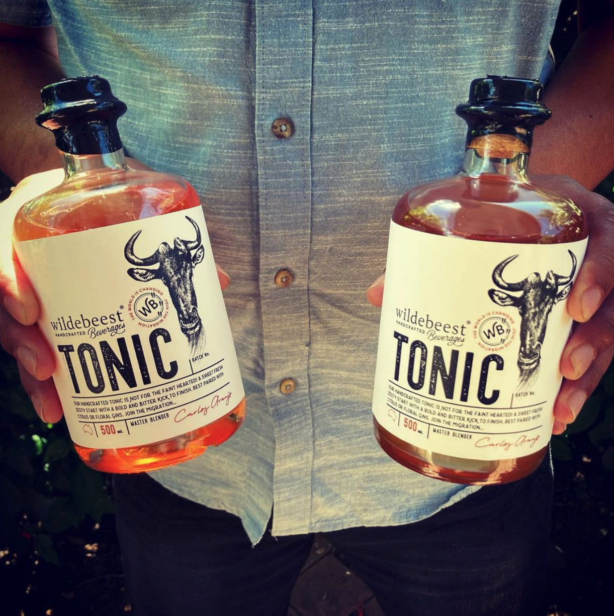 A person wearing a gray button-up shirt holds two bottles of Wilderbeest Tonic, each containing an amber-colored liquid, with both hands.