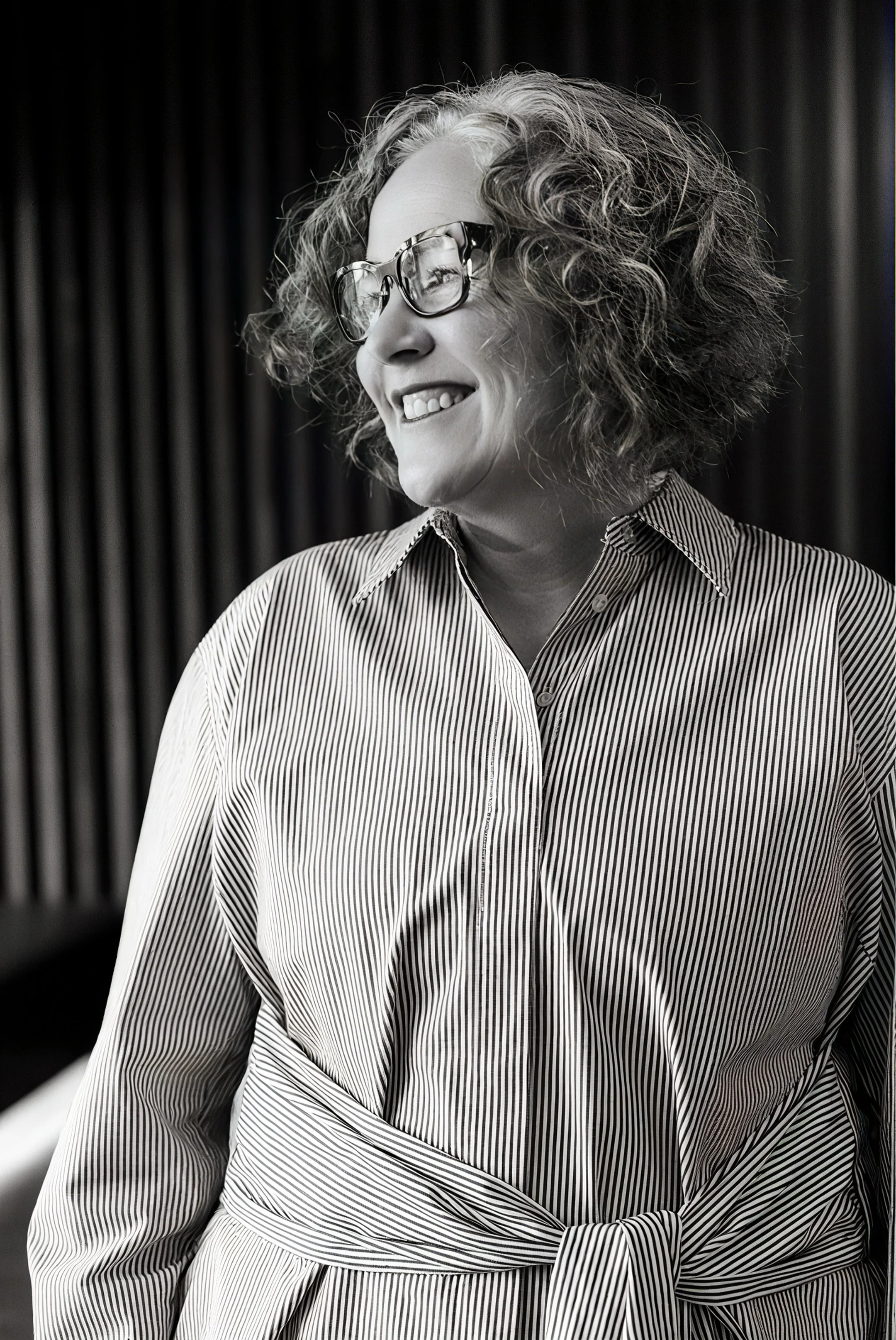 A smiling woman with curly hair and glasses wearing a striped shirt