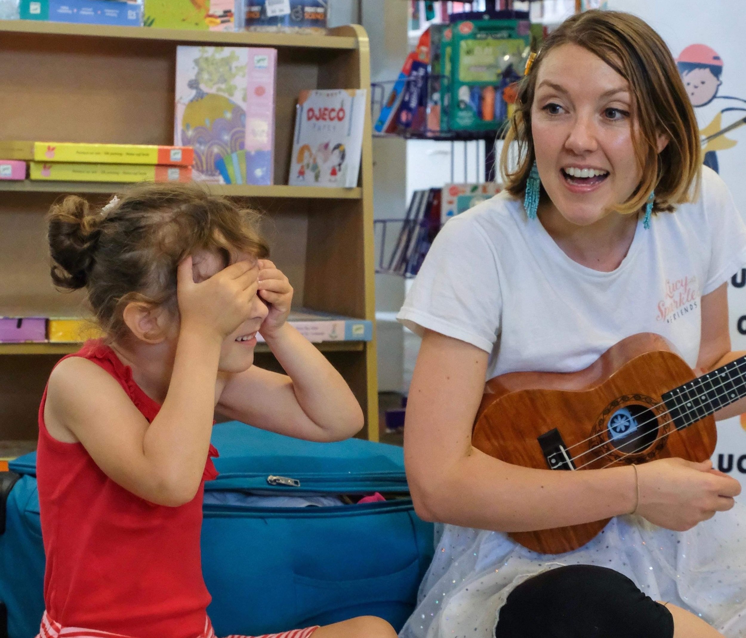 A young girl with curly hair in a bun, wearing a red sleeveless top, covers her eyes and laughs while sitting next to a woman with short red hair, playing a guitar and smiling in a room filled with books and colorful decorations.
