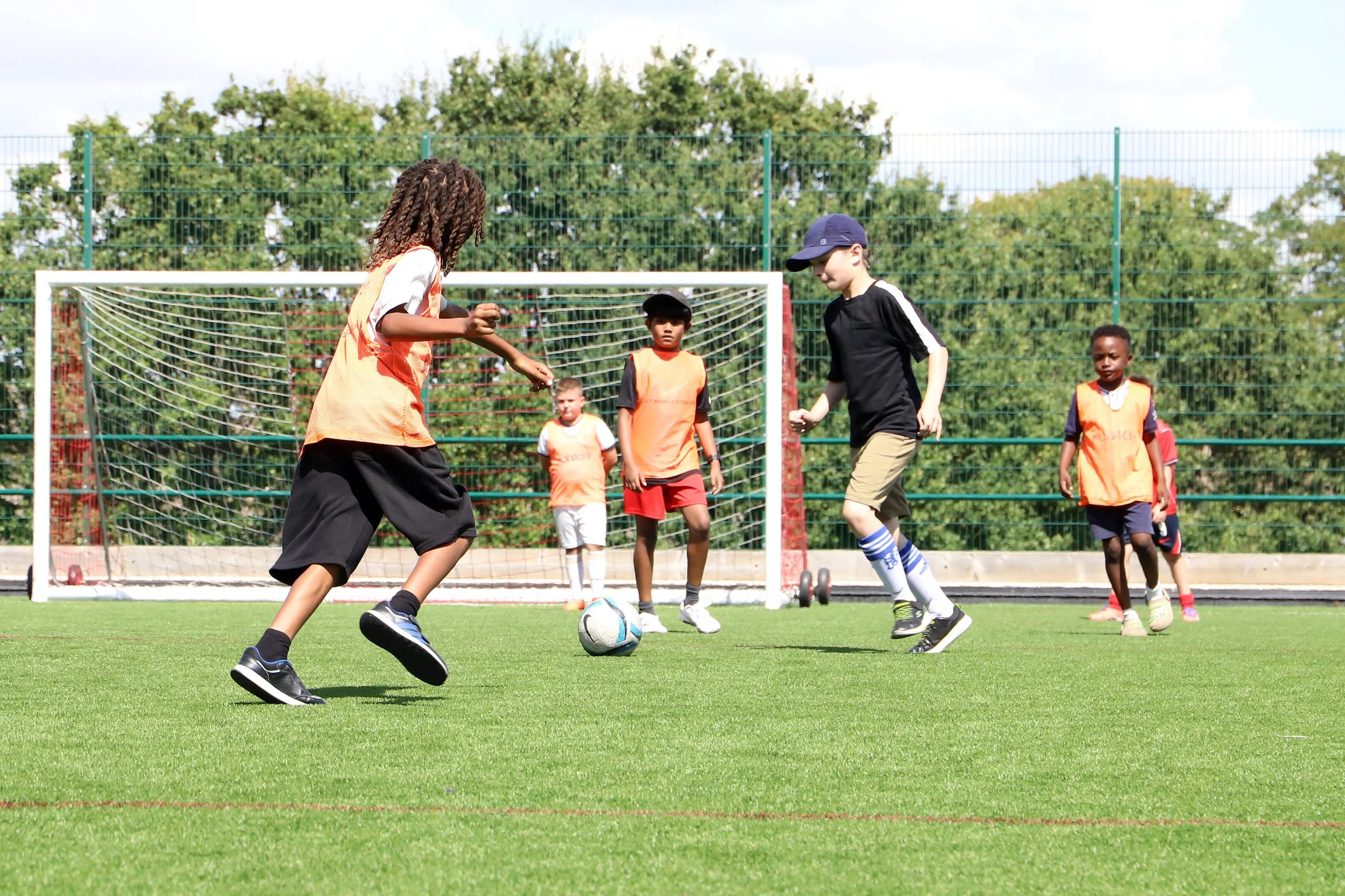 Children playing indoor football on a green artificial turf field, wearing orange bibs and athletic clothing, with a soccer ball in play, surrounded by a fence and trees in the background.