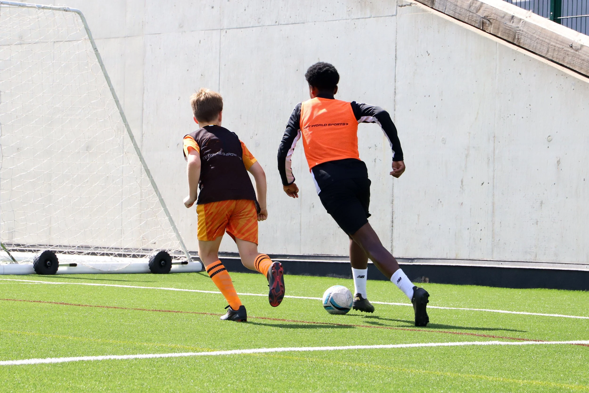 Two boys playing football on a field near a goal, one in orange and black sports attire and the other in a black and red uniform, competing for the ball.