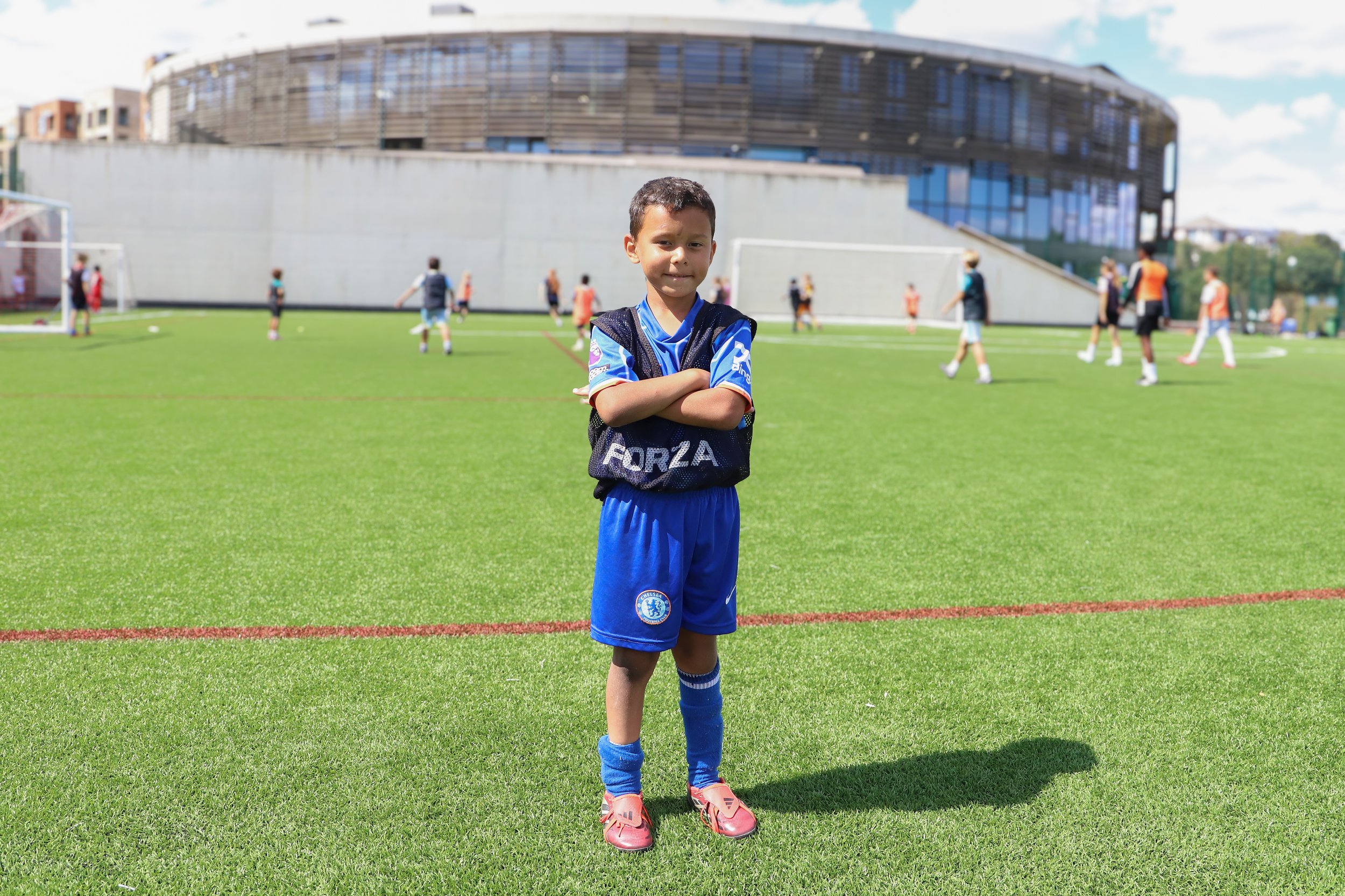 Young boy standing with arms crossed on a football field, wearing a blue soccer uniform and pink cleats, with other children playing soccer in the background.