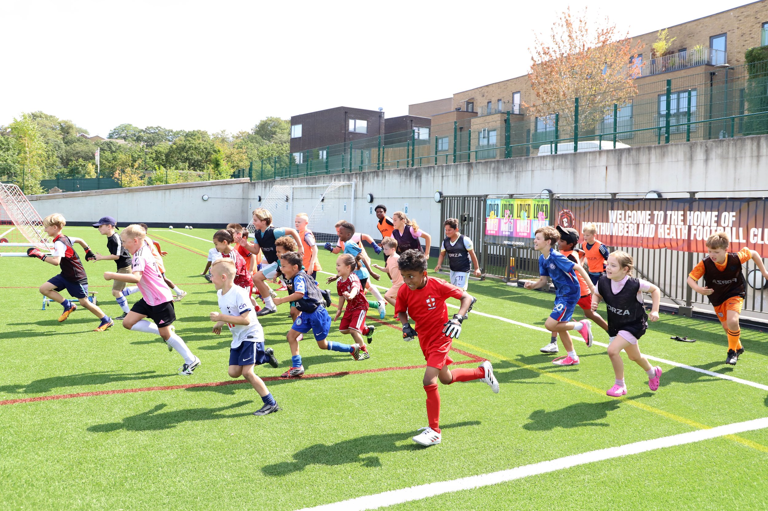 Young children playing football on a green field during daytime, with some wearing colorful sports uniforms and others in casual clothes.