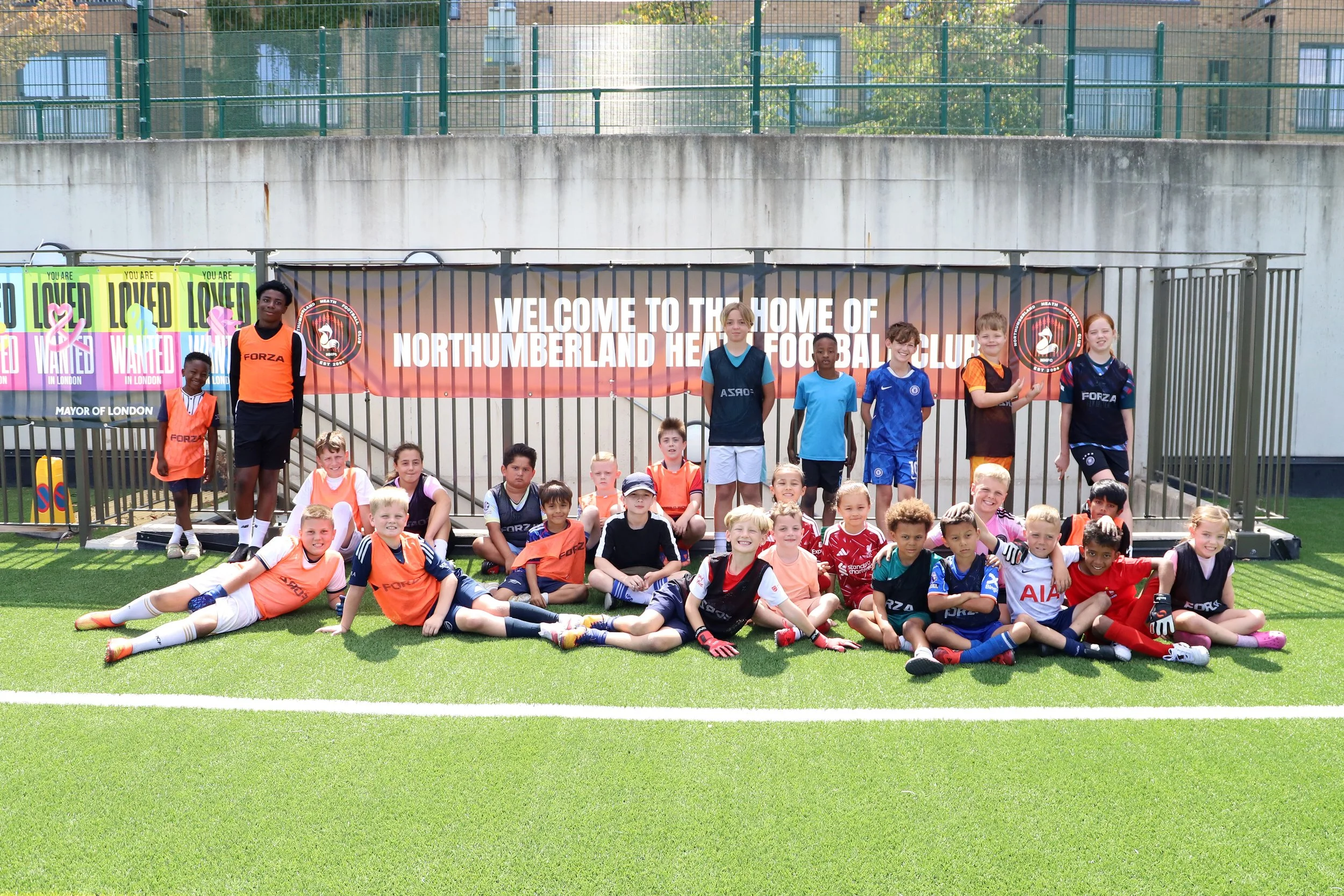 Group of children and teenagers posing for a photo on a football field, with a banner reading 'Welcome to the home of Northumberland Hart Football Club' in the background.